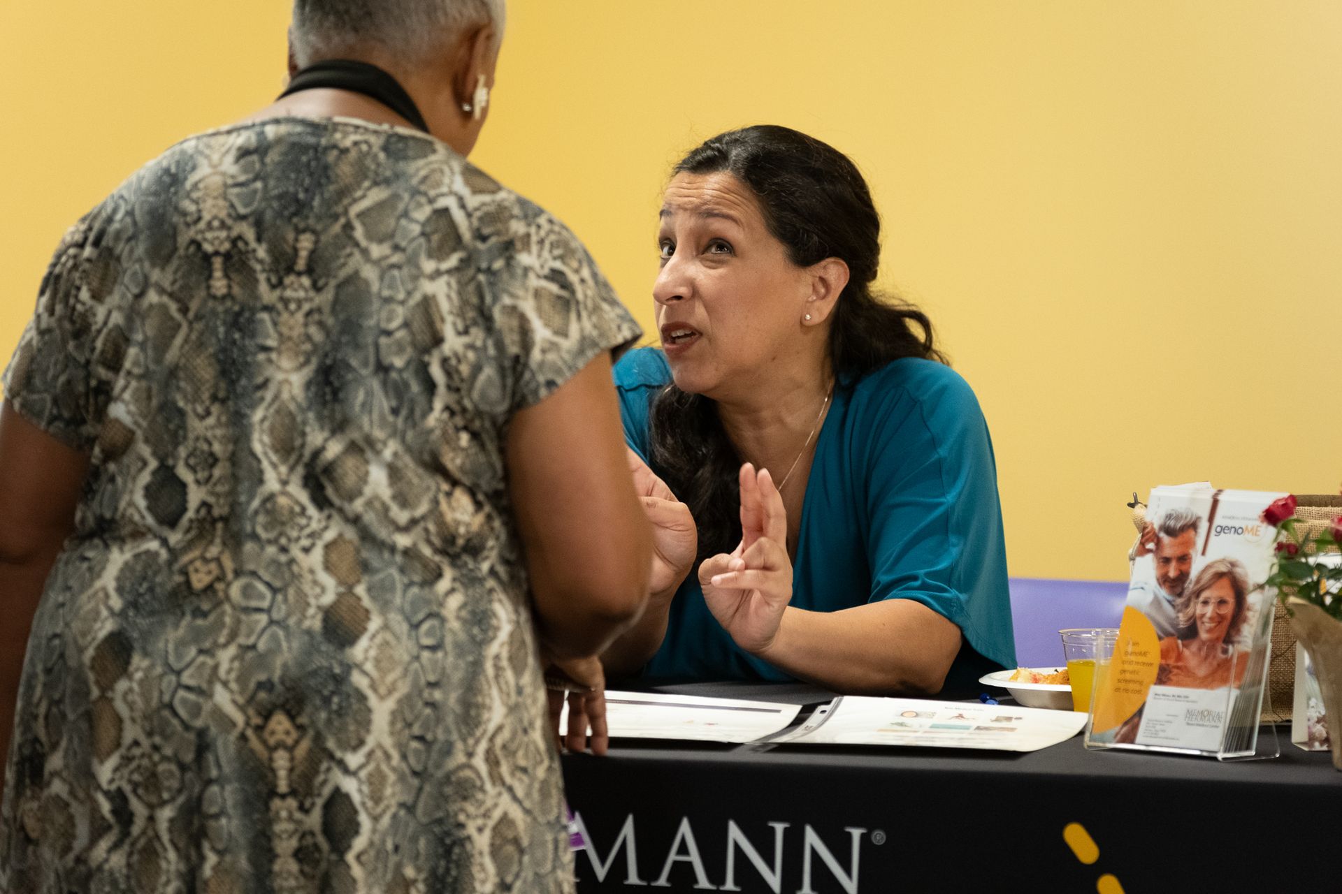 Woman gesturing while speaking to another woman at a table with promotional materials.