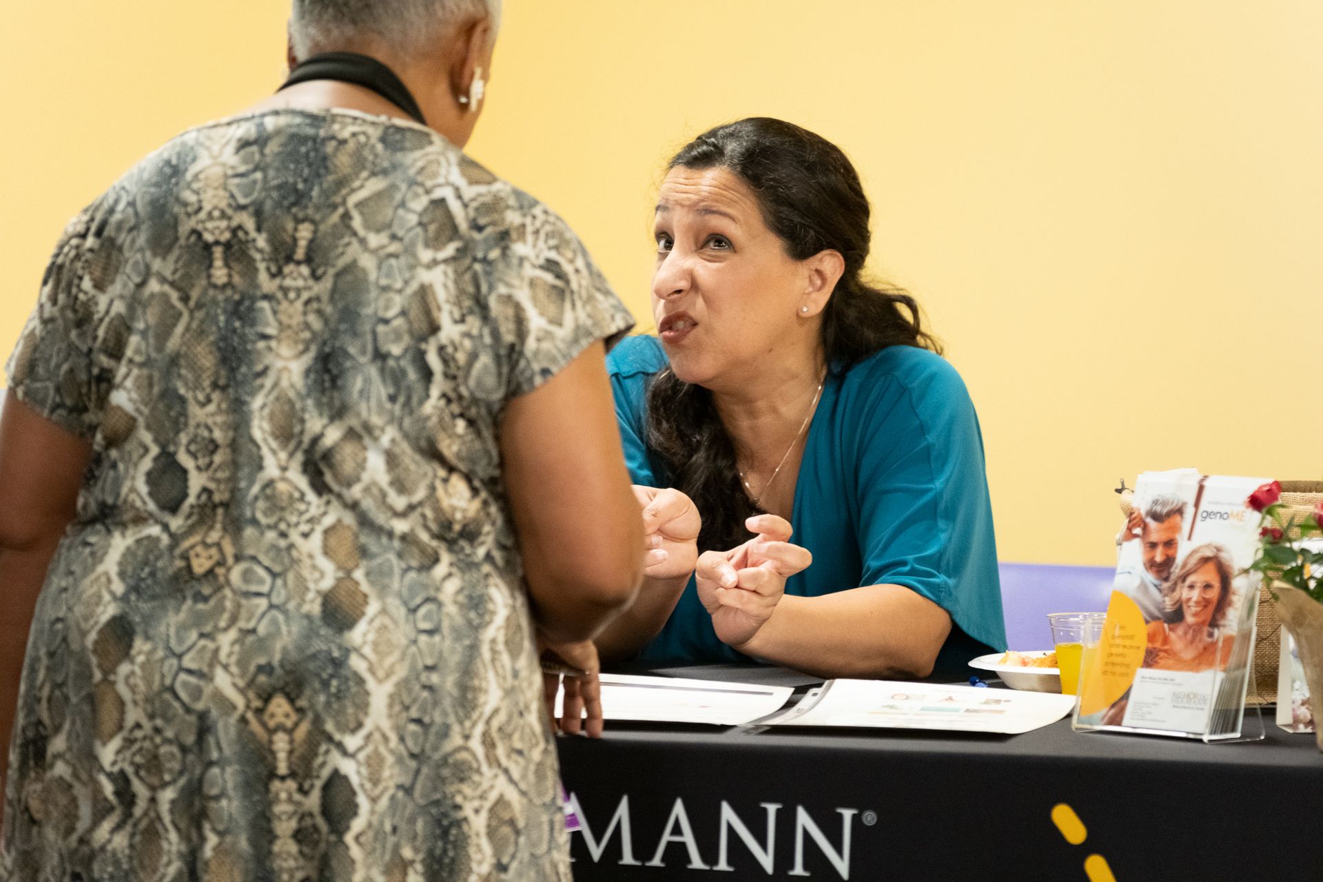 Woman at a table gesturing, talking to another person. The word 