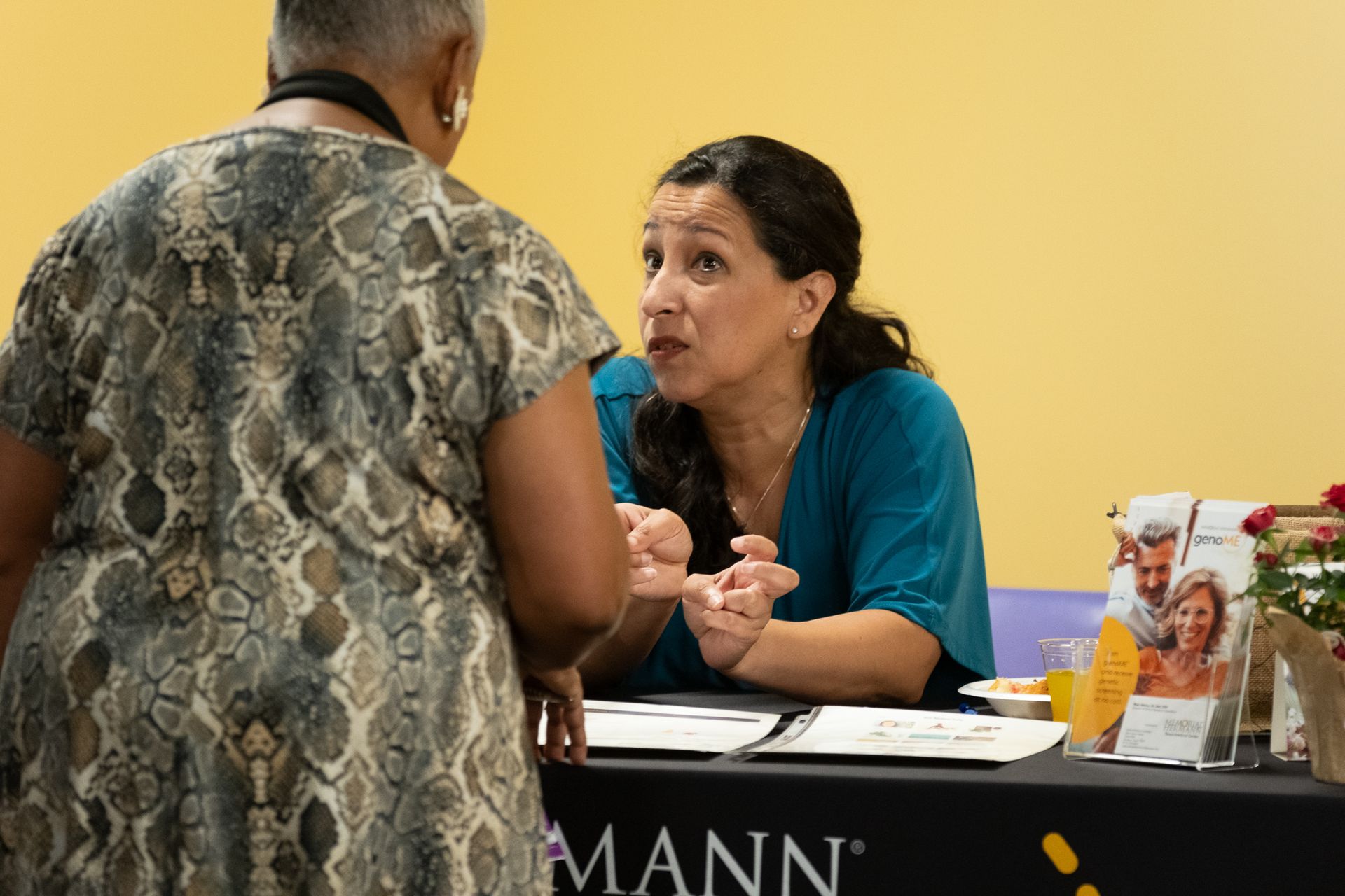 Woman at a table in conversation with another person; brochures visible.