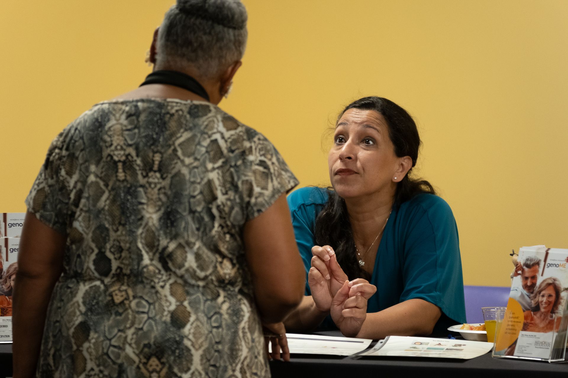 Woman in teal top speaking to another woman in front of a table.