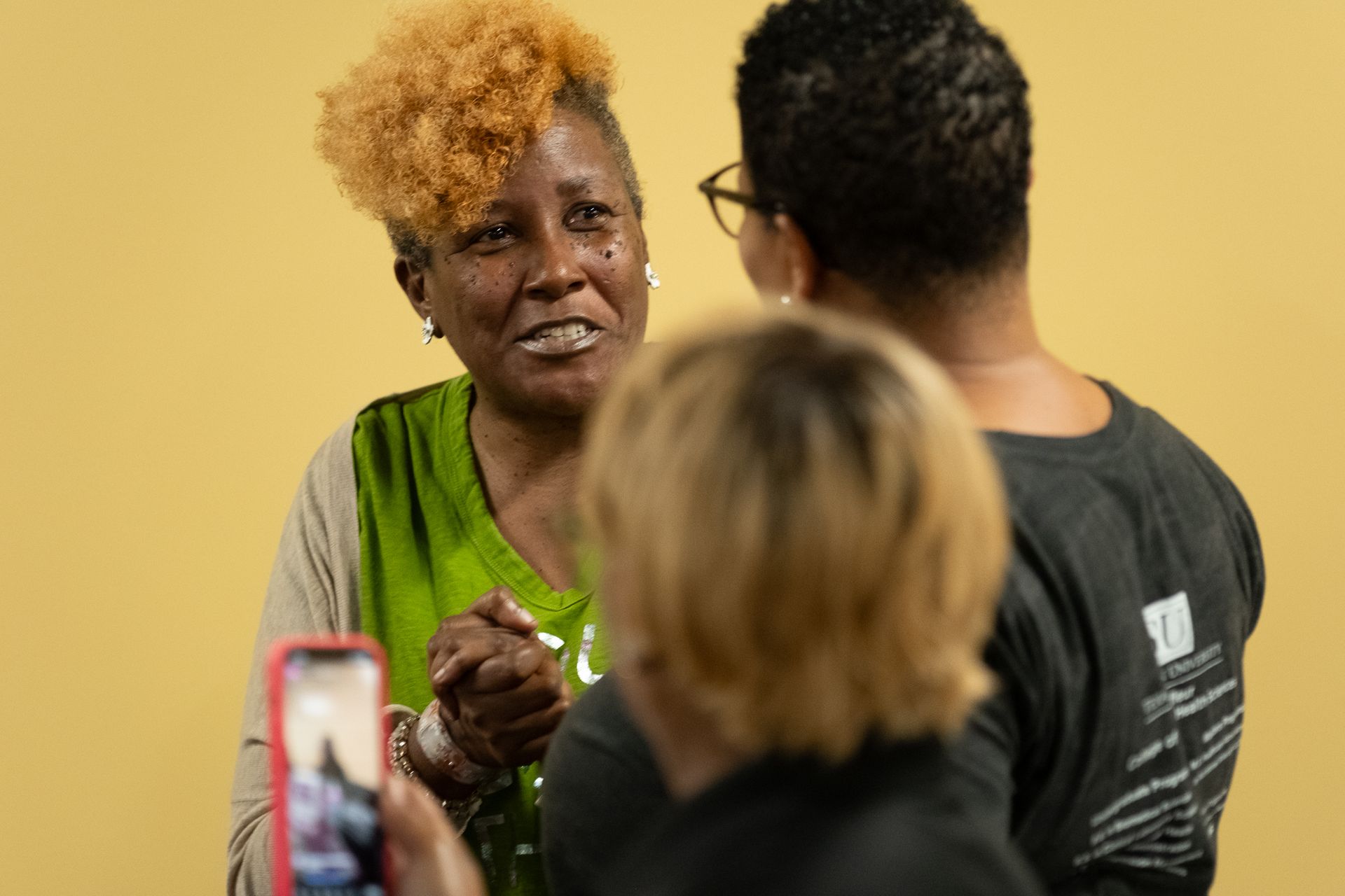 Woman with highlighted hair smiles, speaking to two people. One holds a phone. Yellow wall background.