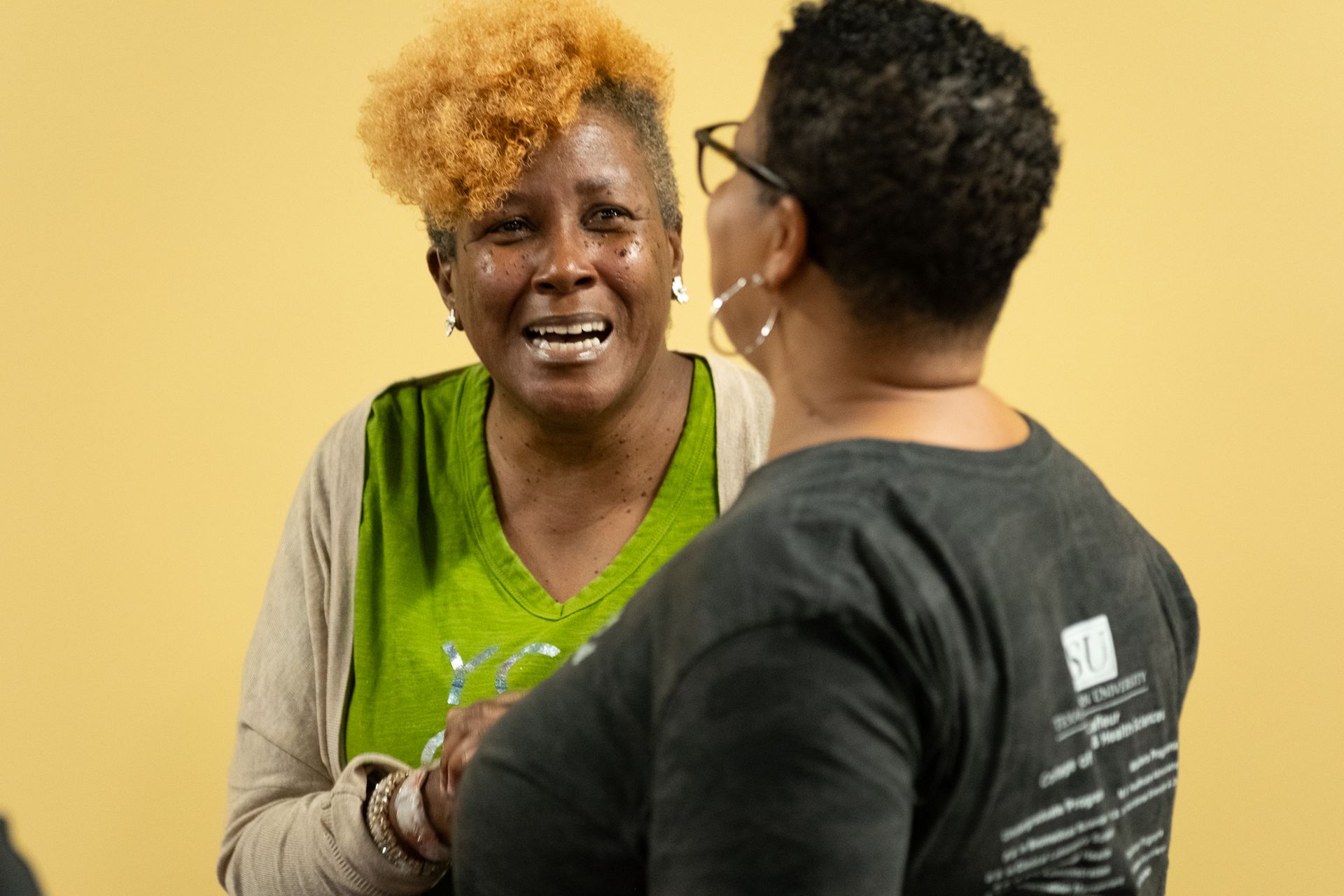 Two people conversing indoors; one with blonde hair smiles.