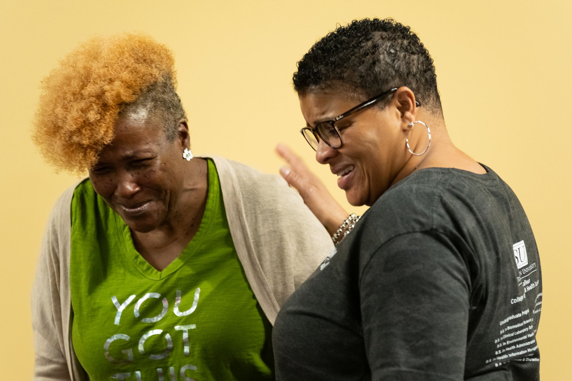 Two people, one with blonde hair, the other with glasses, embracing and possibly crying against a yellow wall.