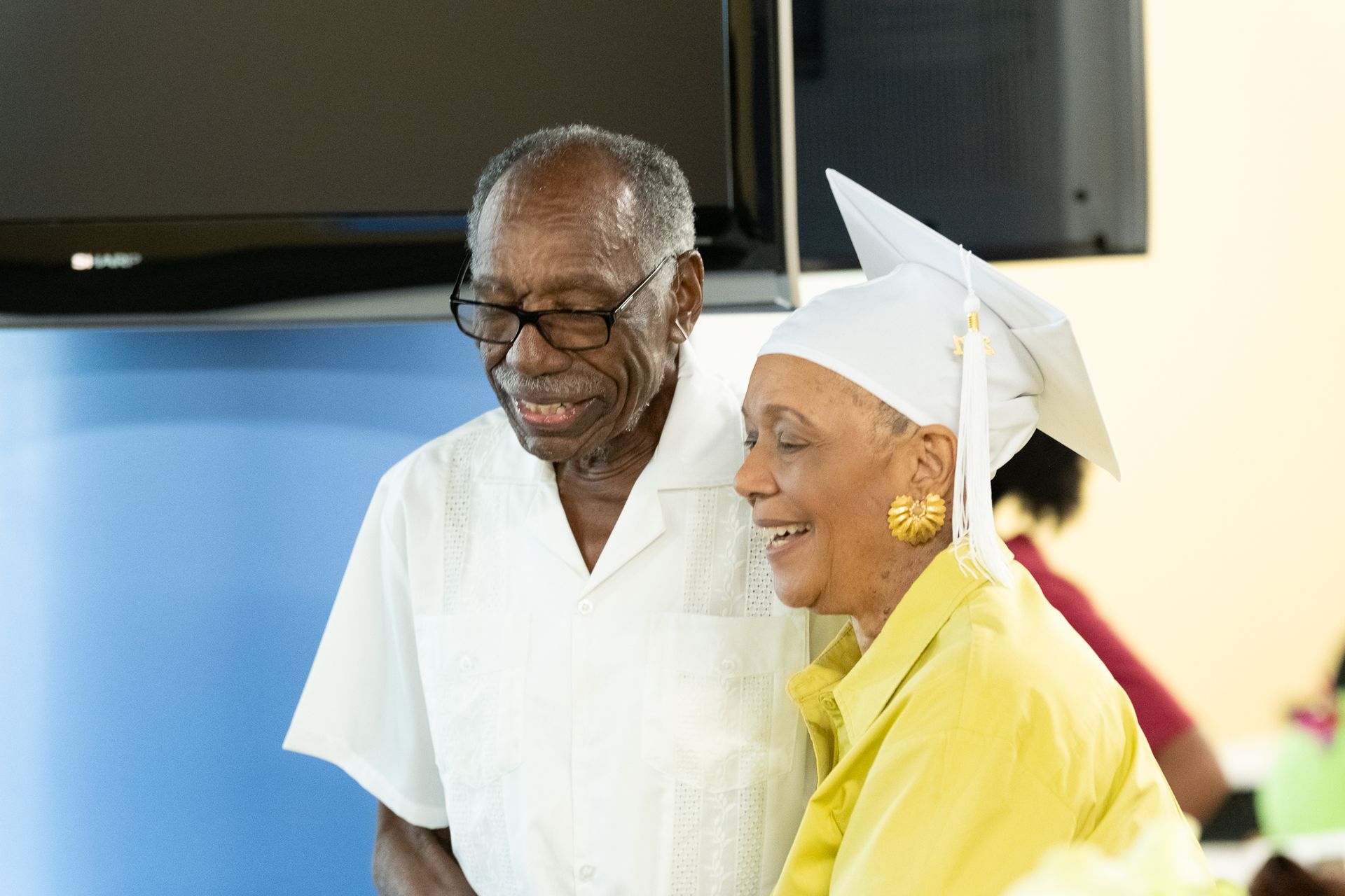 Older couple smiles; woman in graduation cap, man in glasses, indoors.