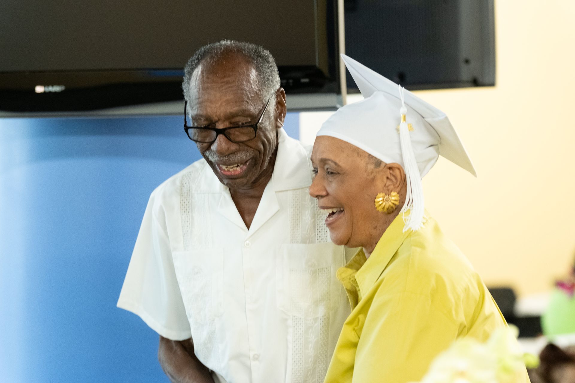 Man and woman smiling, woman in graduation cap and gown, indoors.