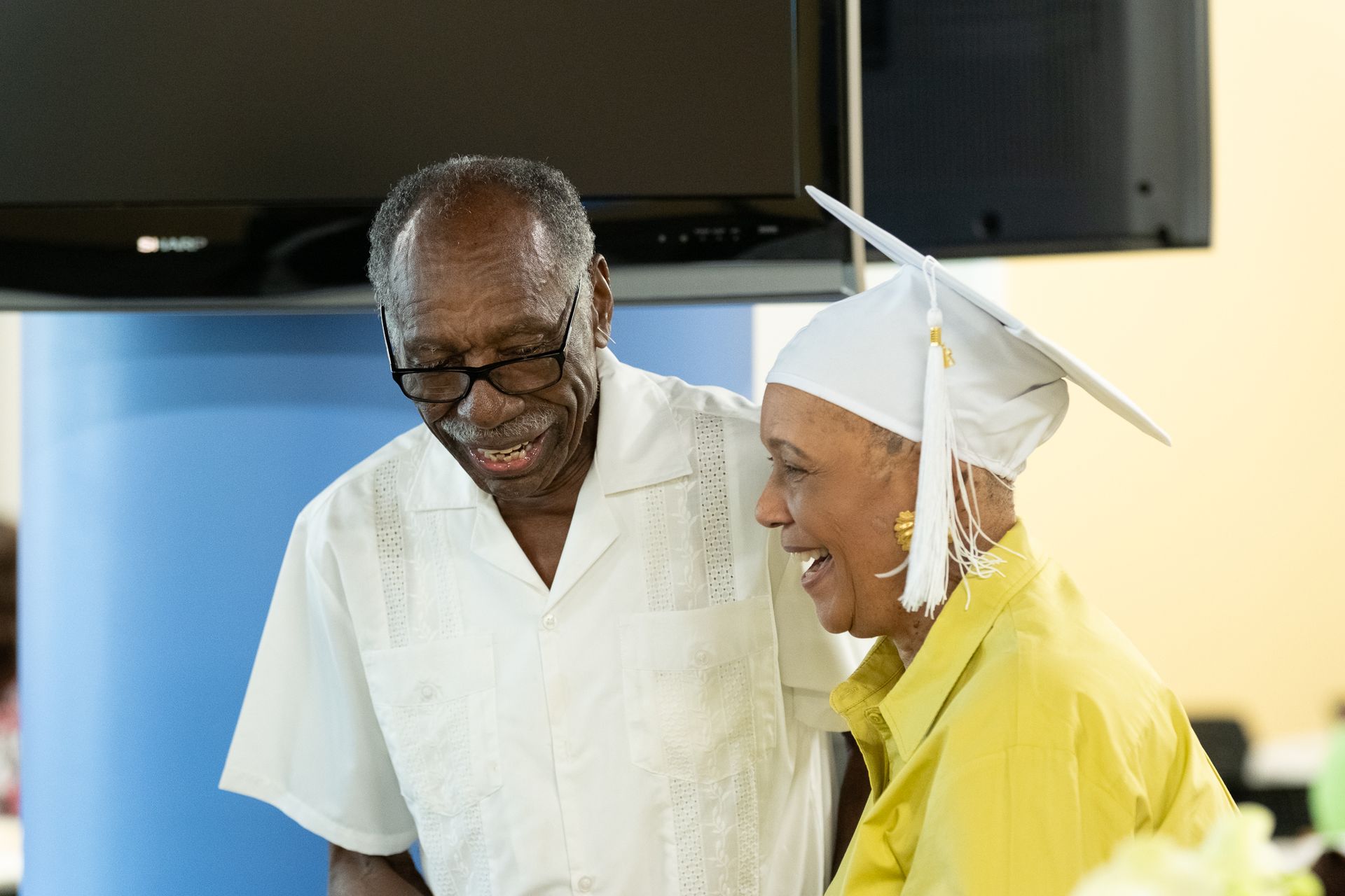 Man and woman smiling; woman wearing graduation cap and yellow shirt, man in glasses and white shirt.