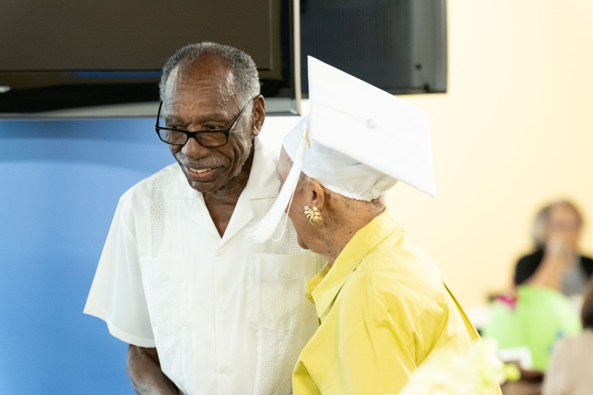 Older person in white shirt talking to person in a yellow top and graduation cap.