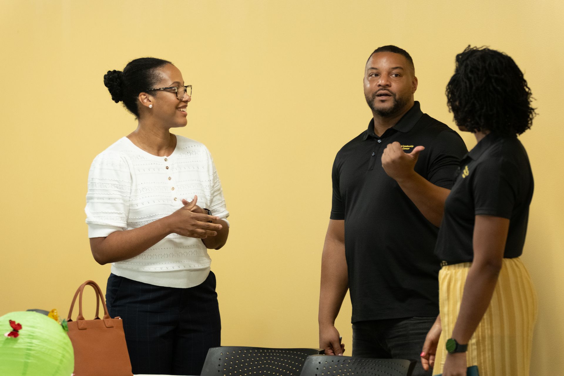 Three people talking in a yellow-walled room; woman in glasses smiles, man points, and a person in yellow skirt listens.