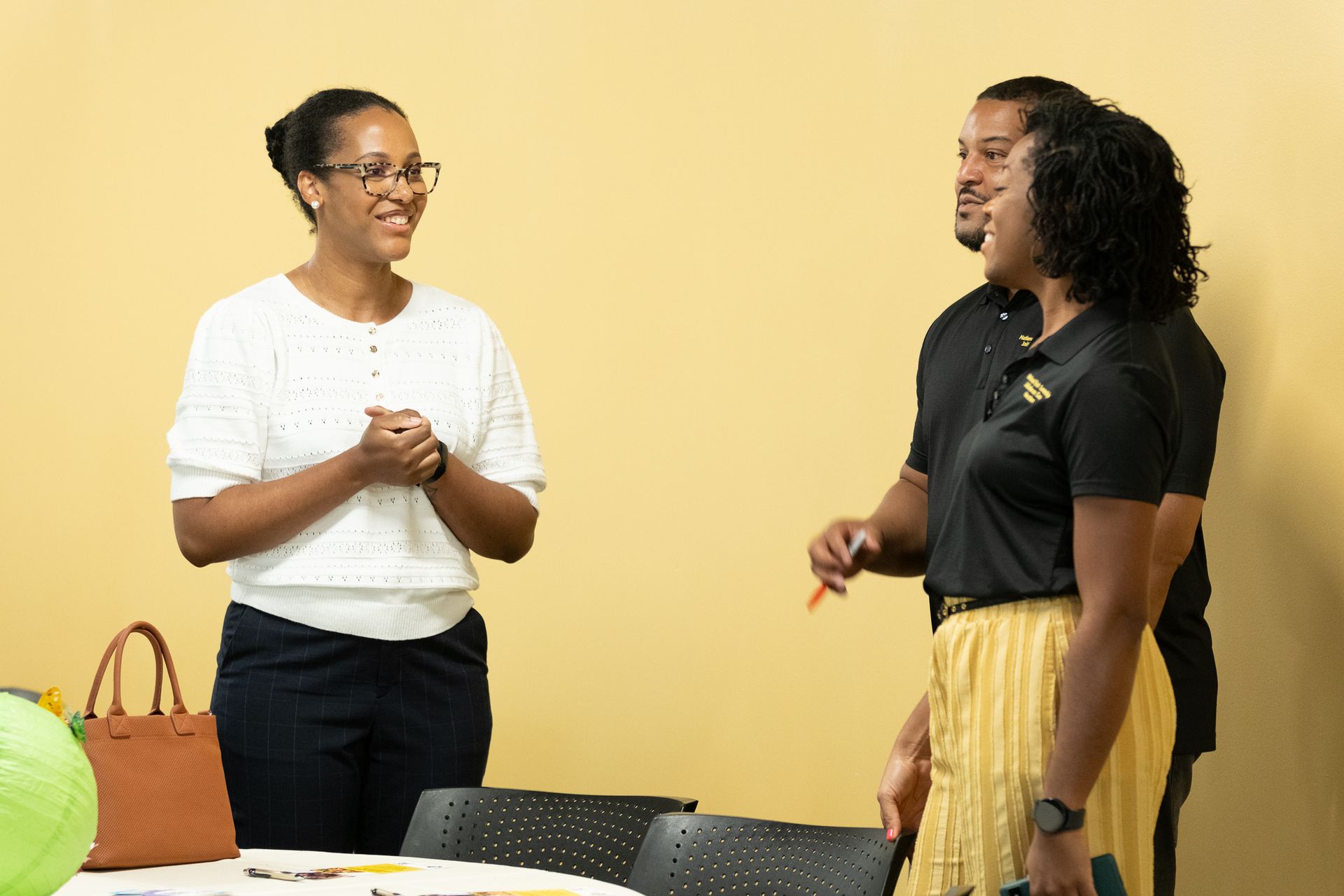 Three people talking and smiling near a table in a room with a light yellow wall.