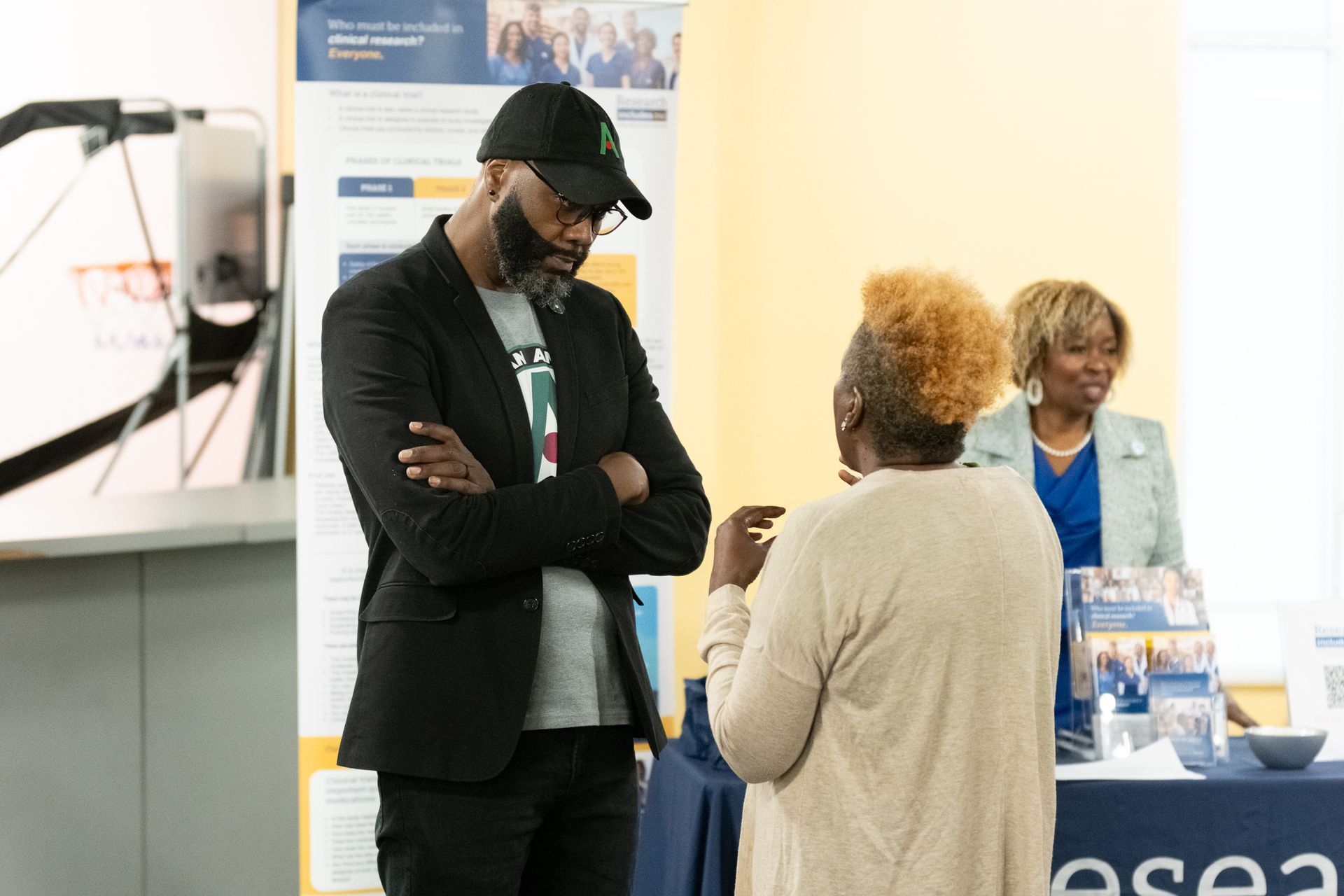 Man in black jacket talks with woman, another woman stands nearby at a display table.