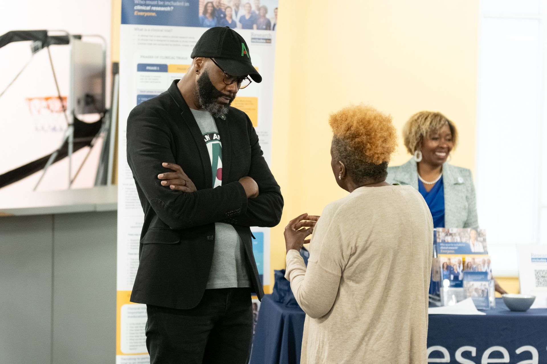 Man in black jacket talks with woman, another woman stands nearby. Tables and poster in background.