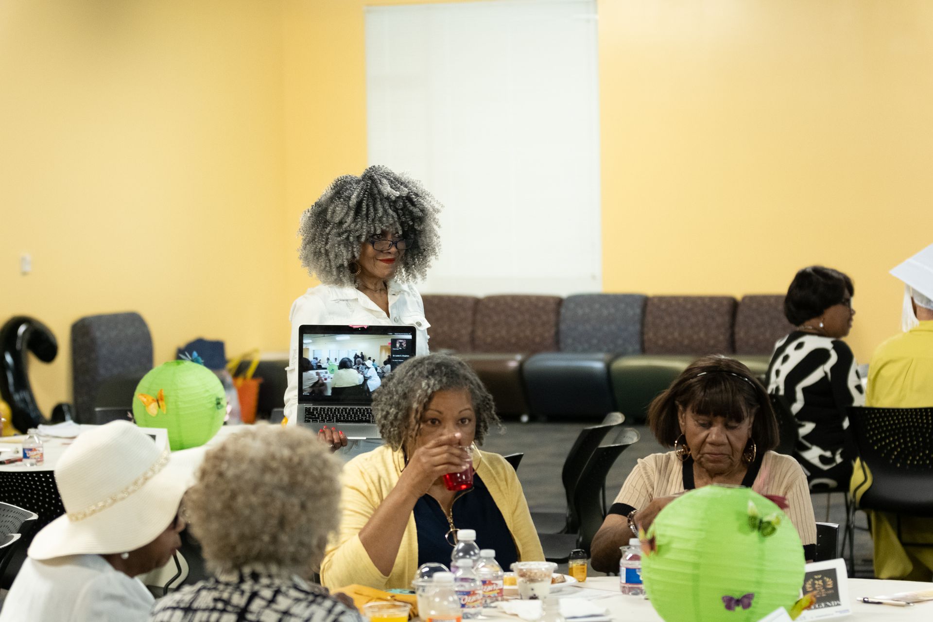 People at a table. Woman with curly hair and a laptop. Green paper lanterns. Yellow walls and seating.