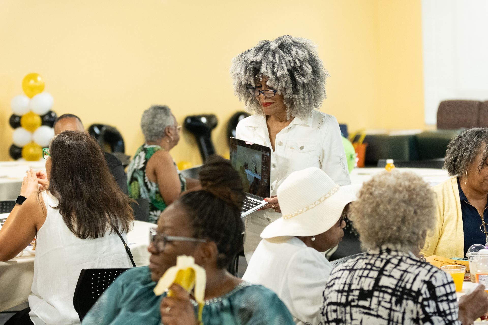 People at a table. A woman with gray hair stands with a laptop. Yellow balloons in the background.
