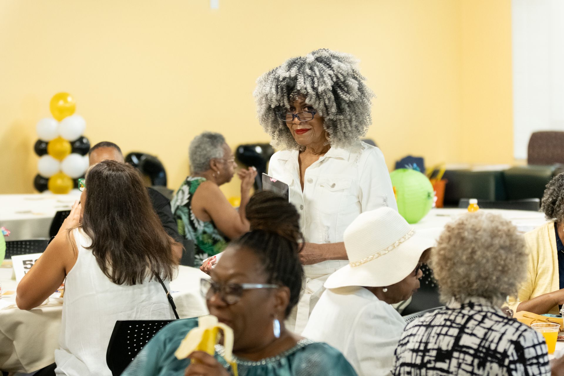 Woman with gray afro, in white outfit, stands at a table in a yellow-walled room, looking at other people. Balloons nearby.