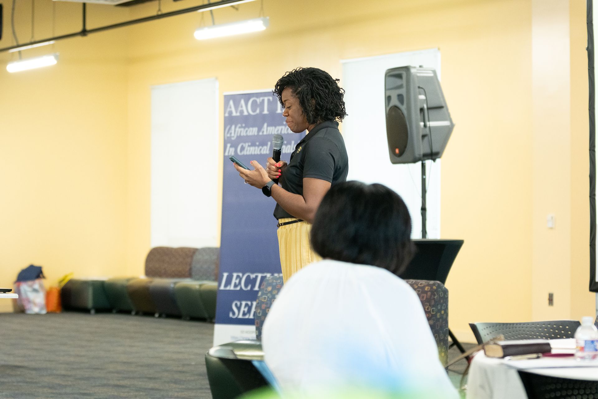 Woman speaking into microphone at a conference; AACT banner visible.