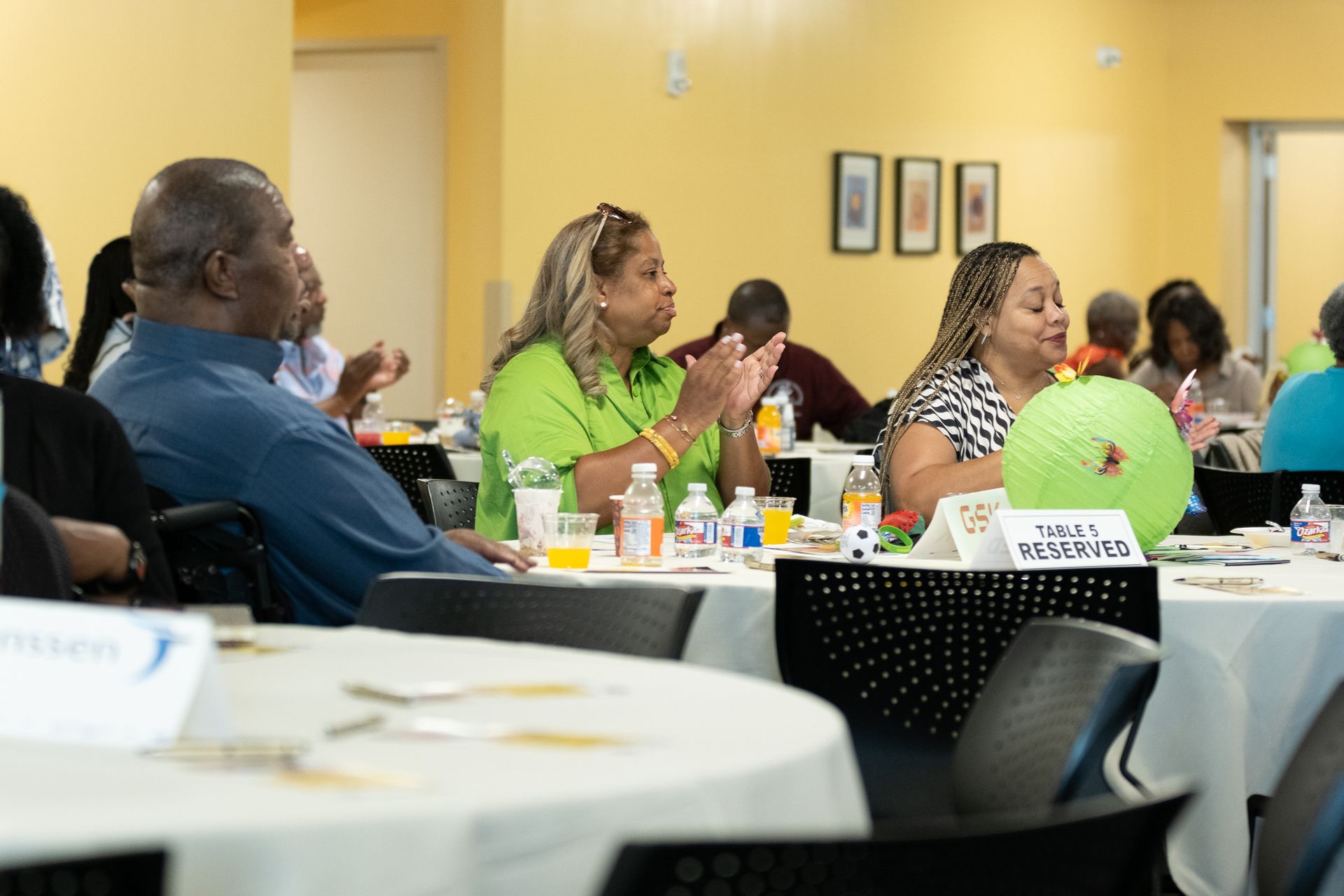 People at tables in a room, clapping, with drinks and decorations on tables.