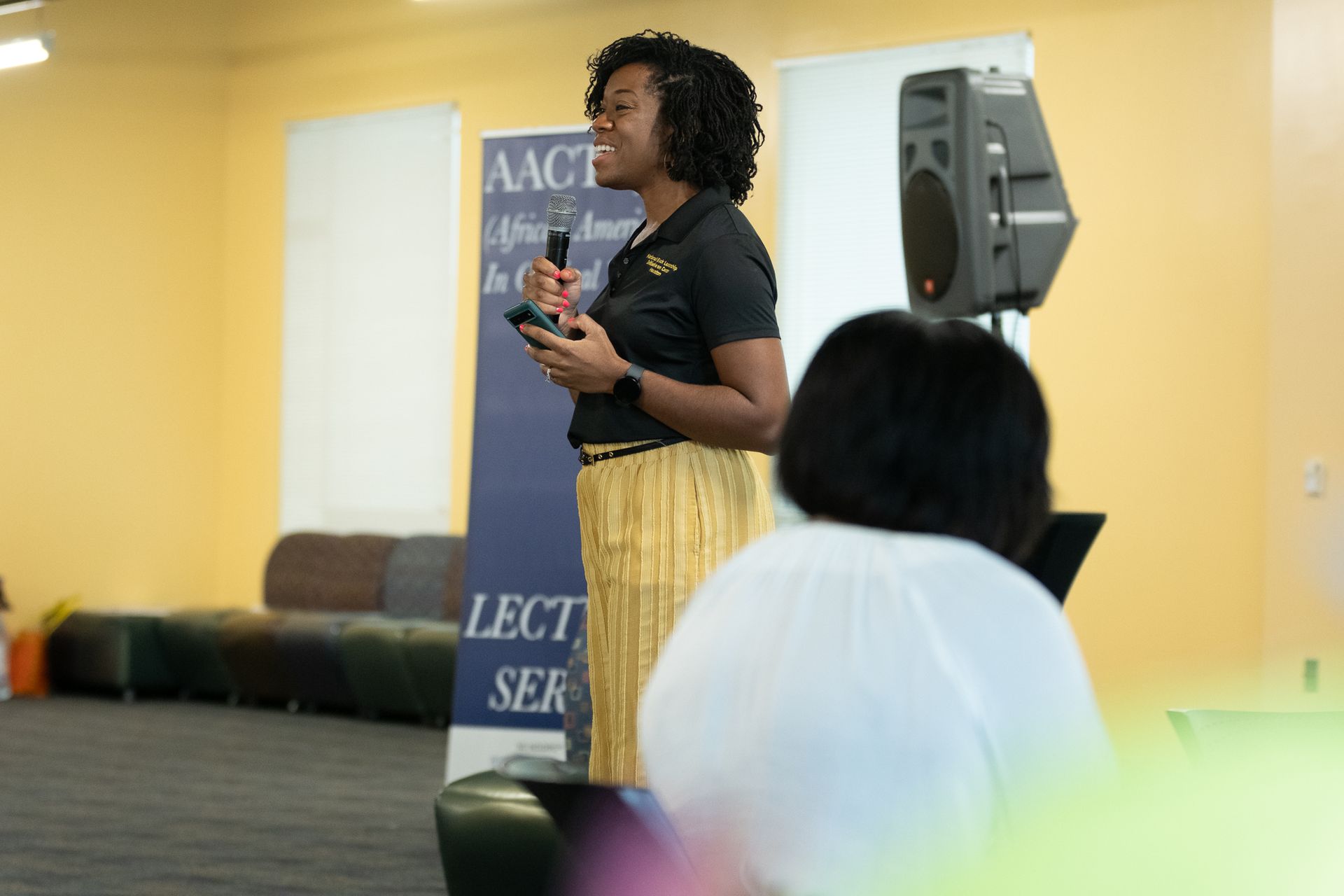 Woman speaking at a lecture, holding microphone. Yellow skirt, black shirt. AACT banner and speaker in the background.