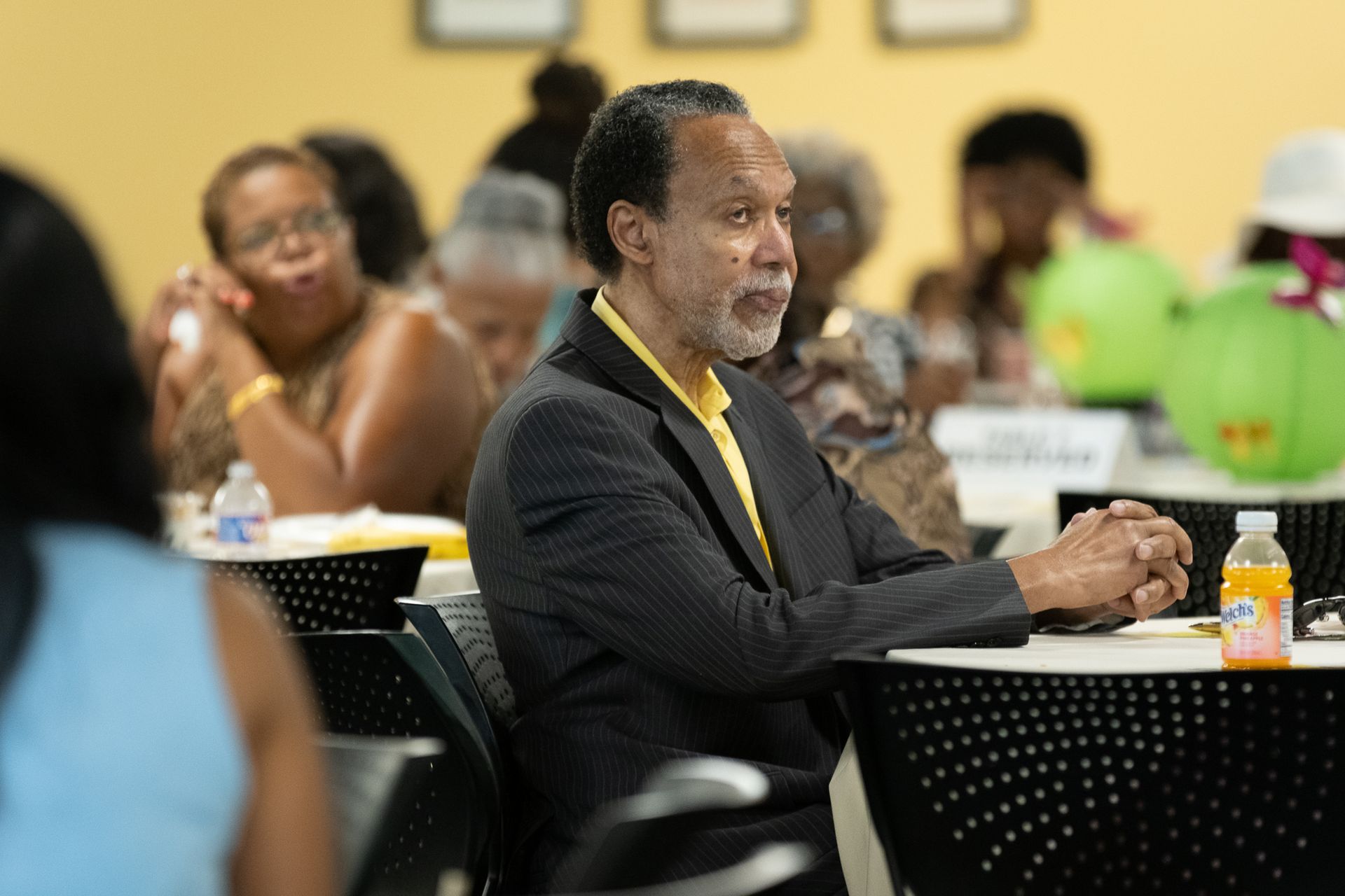 Man in a suit at a table, looking to the side, at an event. Other people are seated at tables in the background.