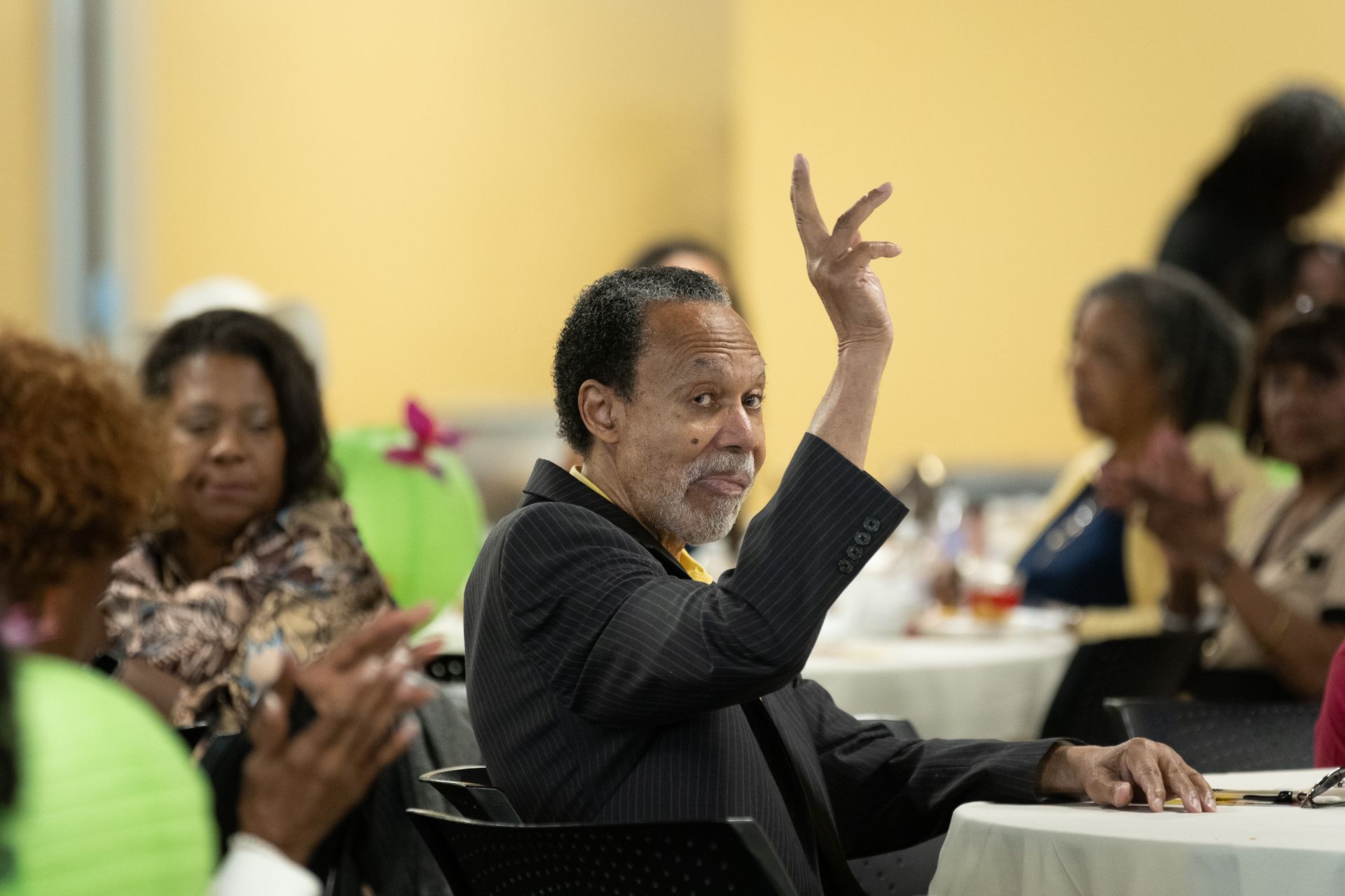 Man in a suit raises hand with a gesture, smiling. Other people seated at tables, applauding. Interior with yellow wall.