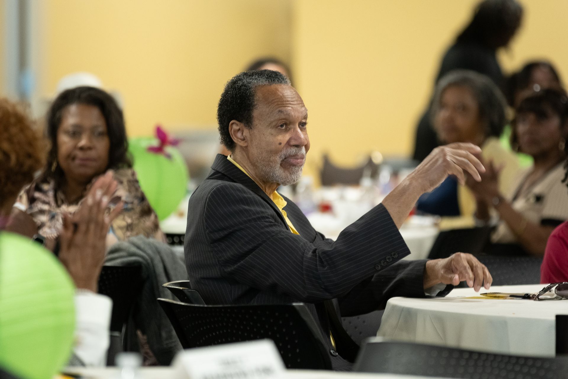 Man at a table gestures while others clap in a room with light green and yellow walls.