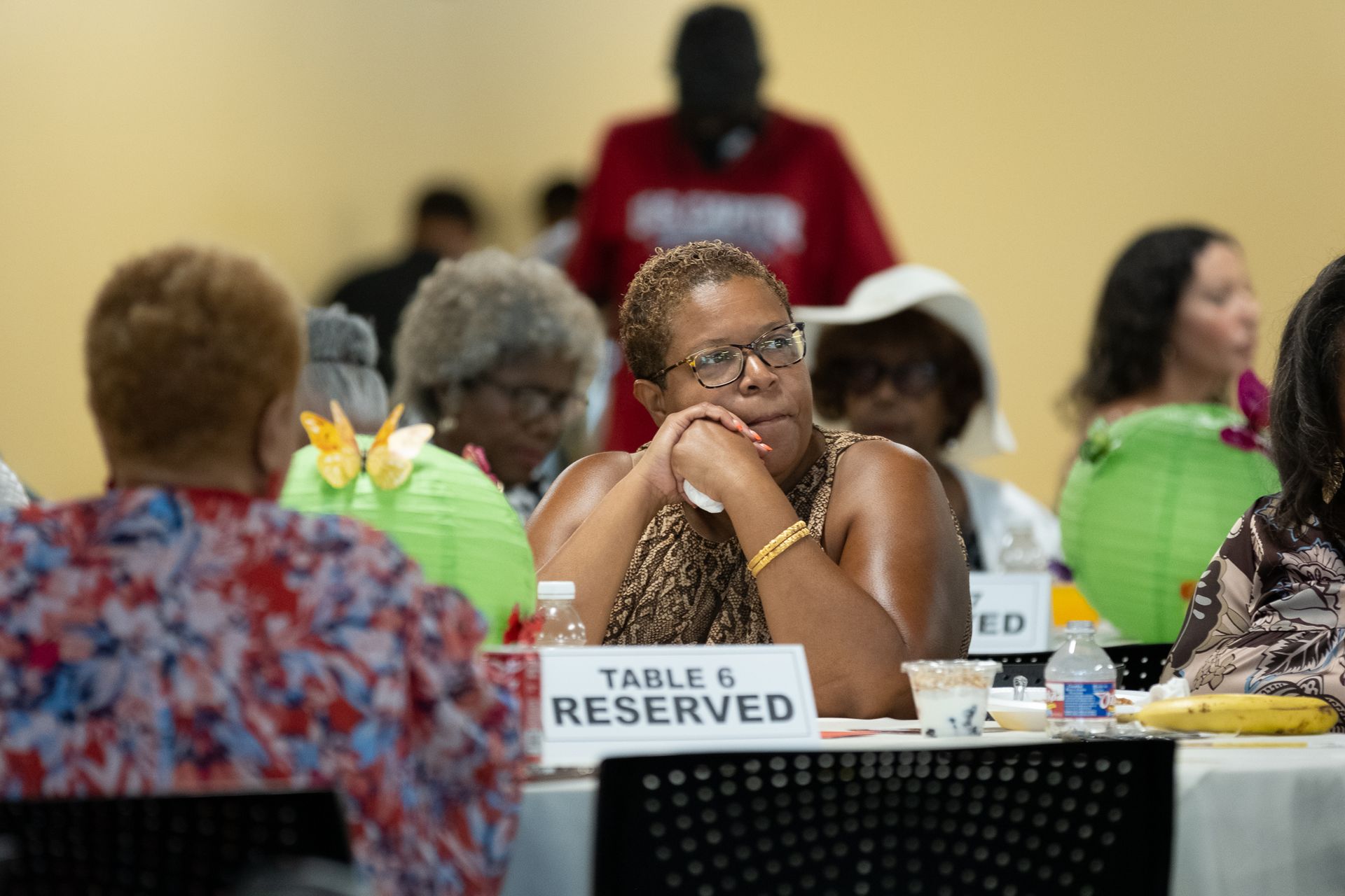 Woman at a reserved table in a meeting, resting chin on hands. Other people at tables in a brightly lit room.