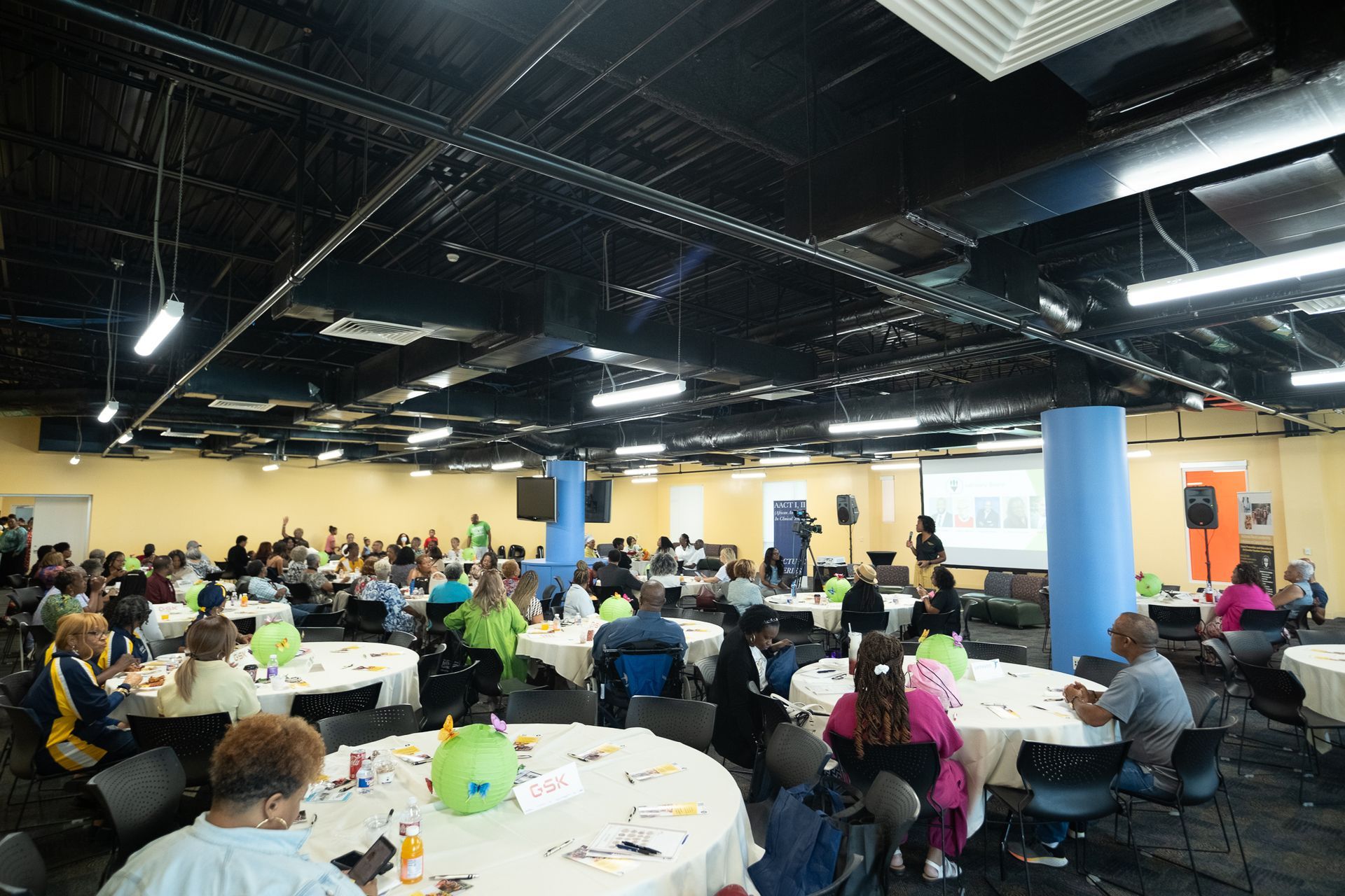 Large room with people at round tables, attending an event. Bright ceiling, blue pillars, and projected screen.