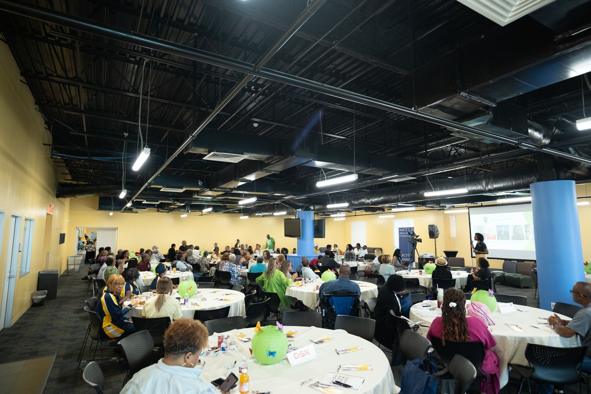 Large room with people at round tables, attending an event. Bright colors, projector screen, blue support beams.
