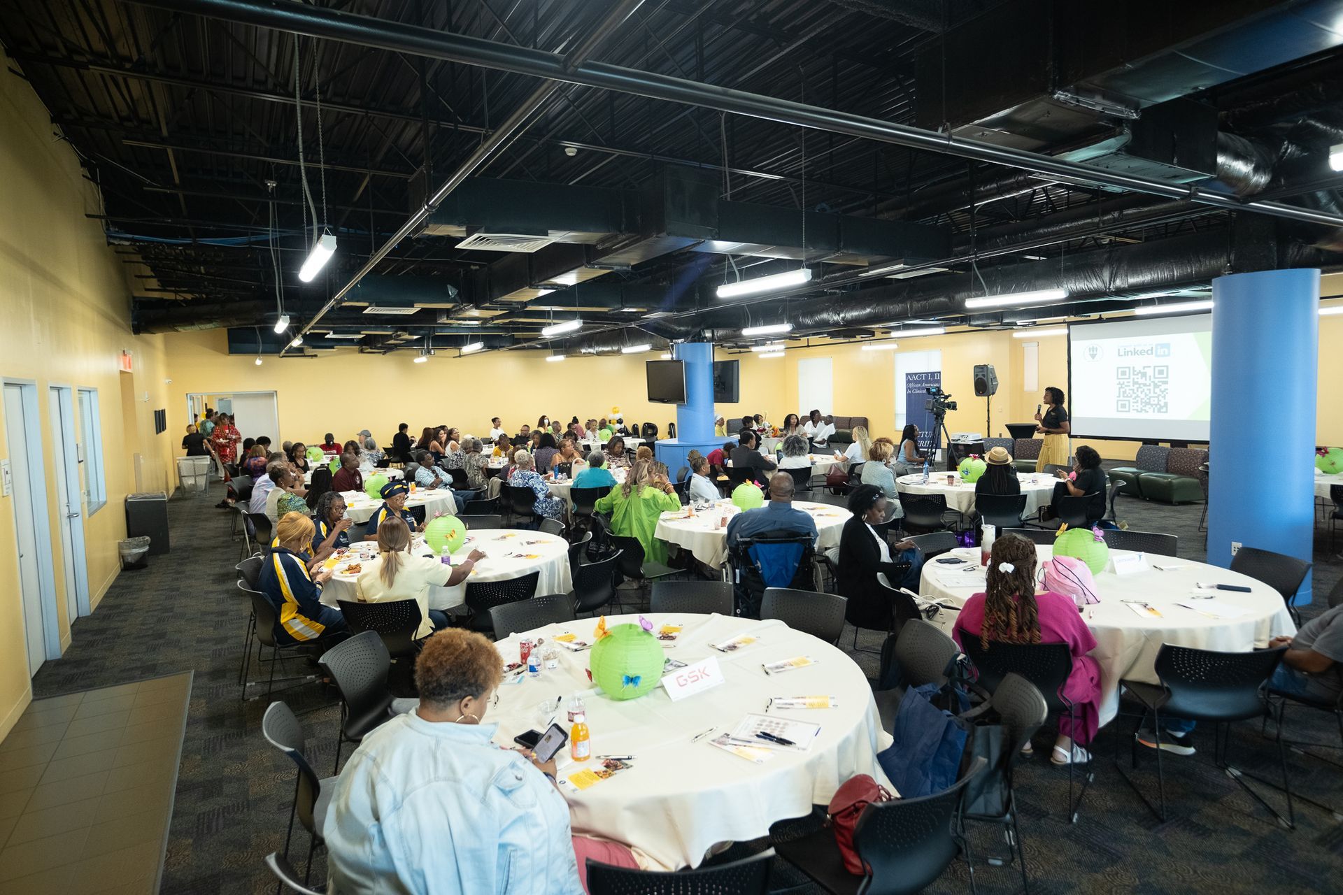 Large room with tables and chairs, filled with people attending an event. A screen displays text.