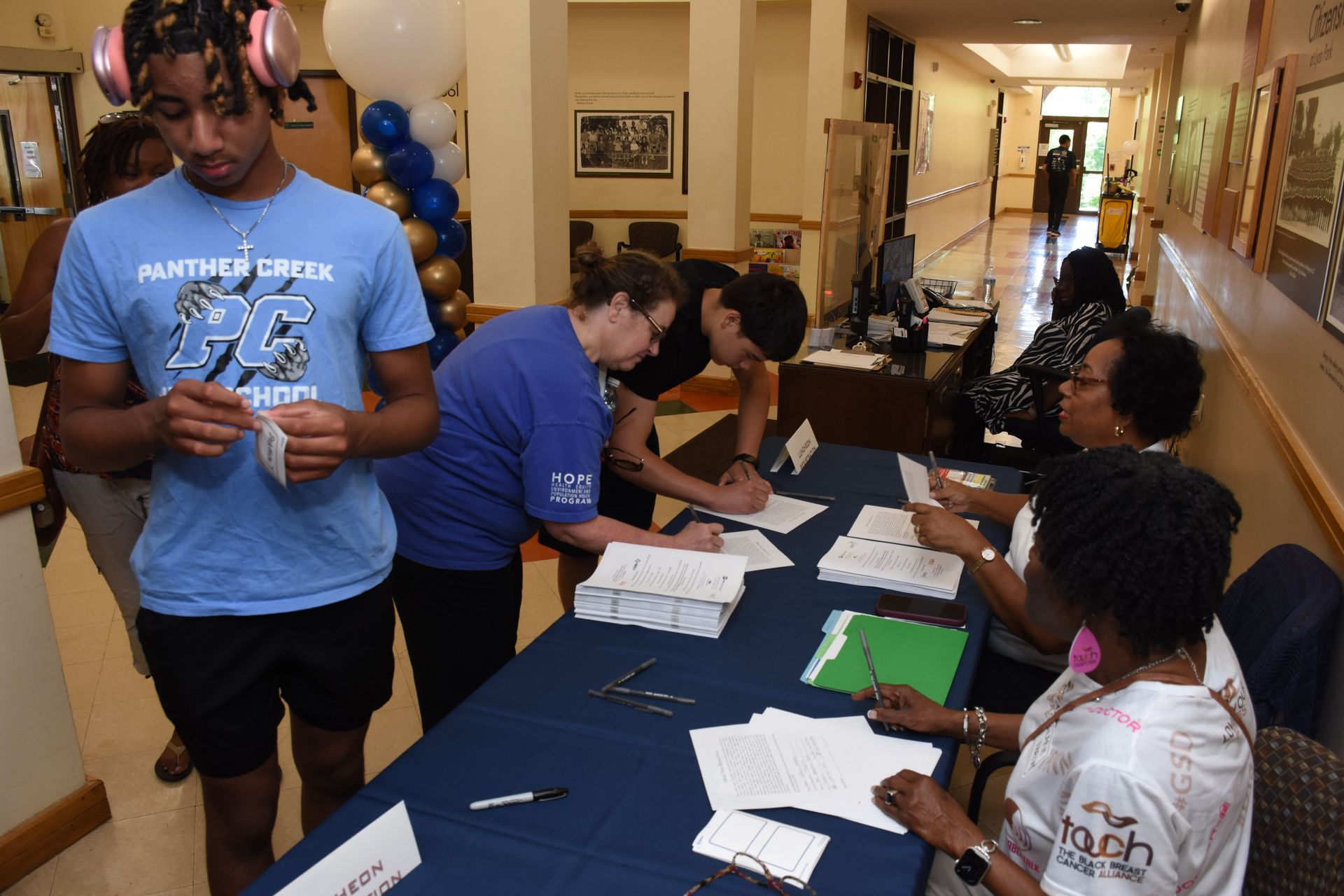 People at a table signing papers in a hallway, balloons in background.