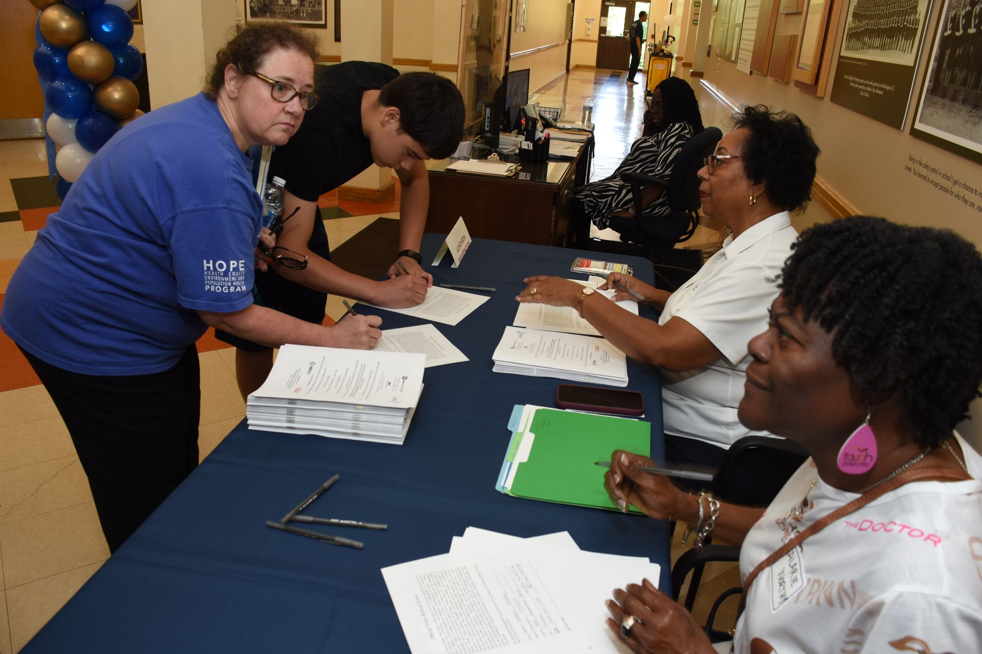 People signing papers at a table with volunteers. Balloons in the background.