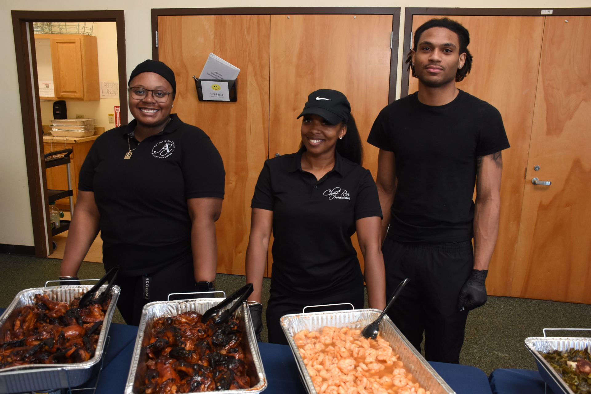 Three people stand behind food trays at an event. They are in black shirts and smiling, next to various dishes.