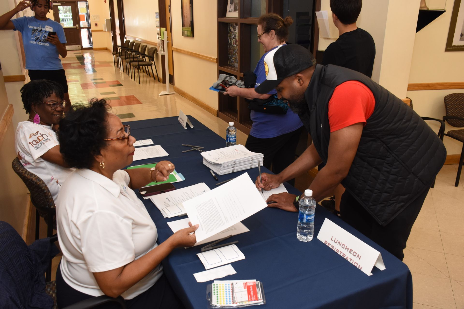 People registering at a table, likely for an event. One man signs a paper, others assist. Indoors.