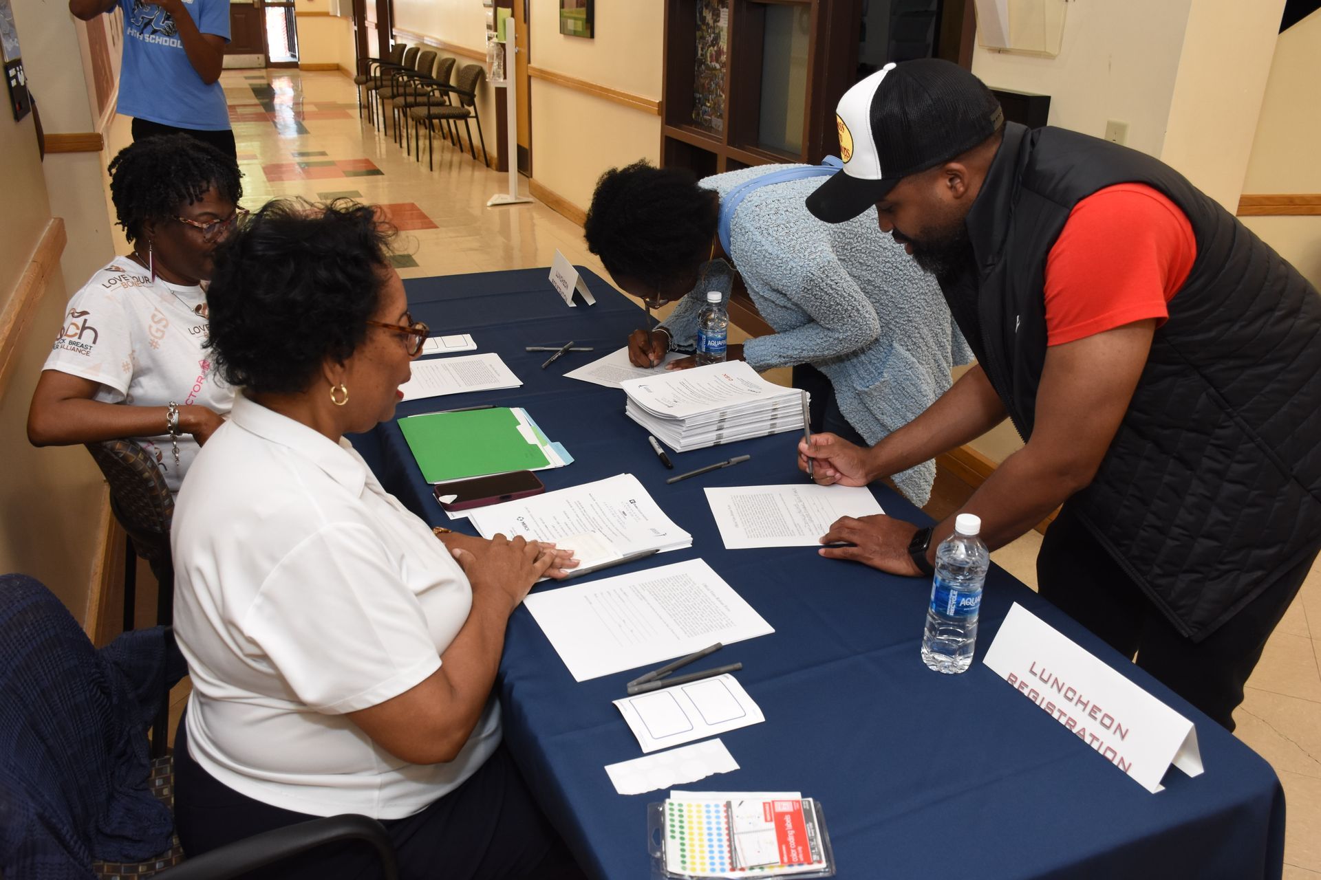 People signing documents at a table. Papers and a water bottle present. Indoor setting.