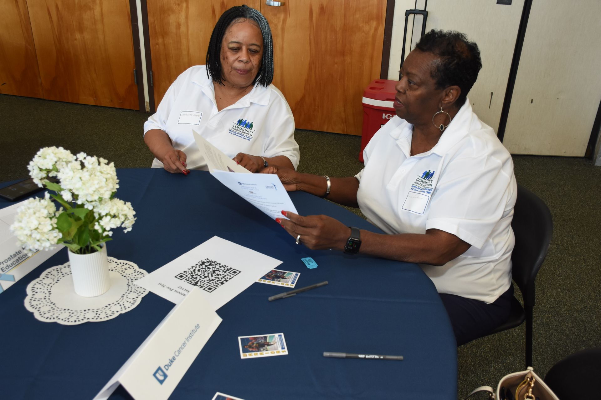 Two women seated at a table, reviewing papers. White shirts, blue tablecloth, flowers in vase.