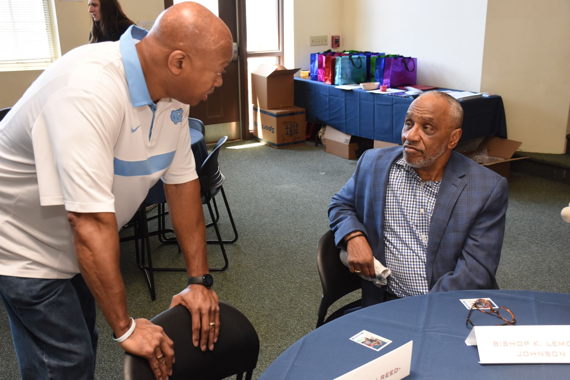 Two men talking at a table in a room. One stands, wearing a light blue shirt; the other sits in a blazer.