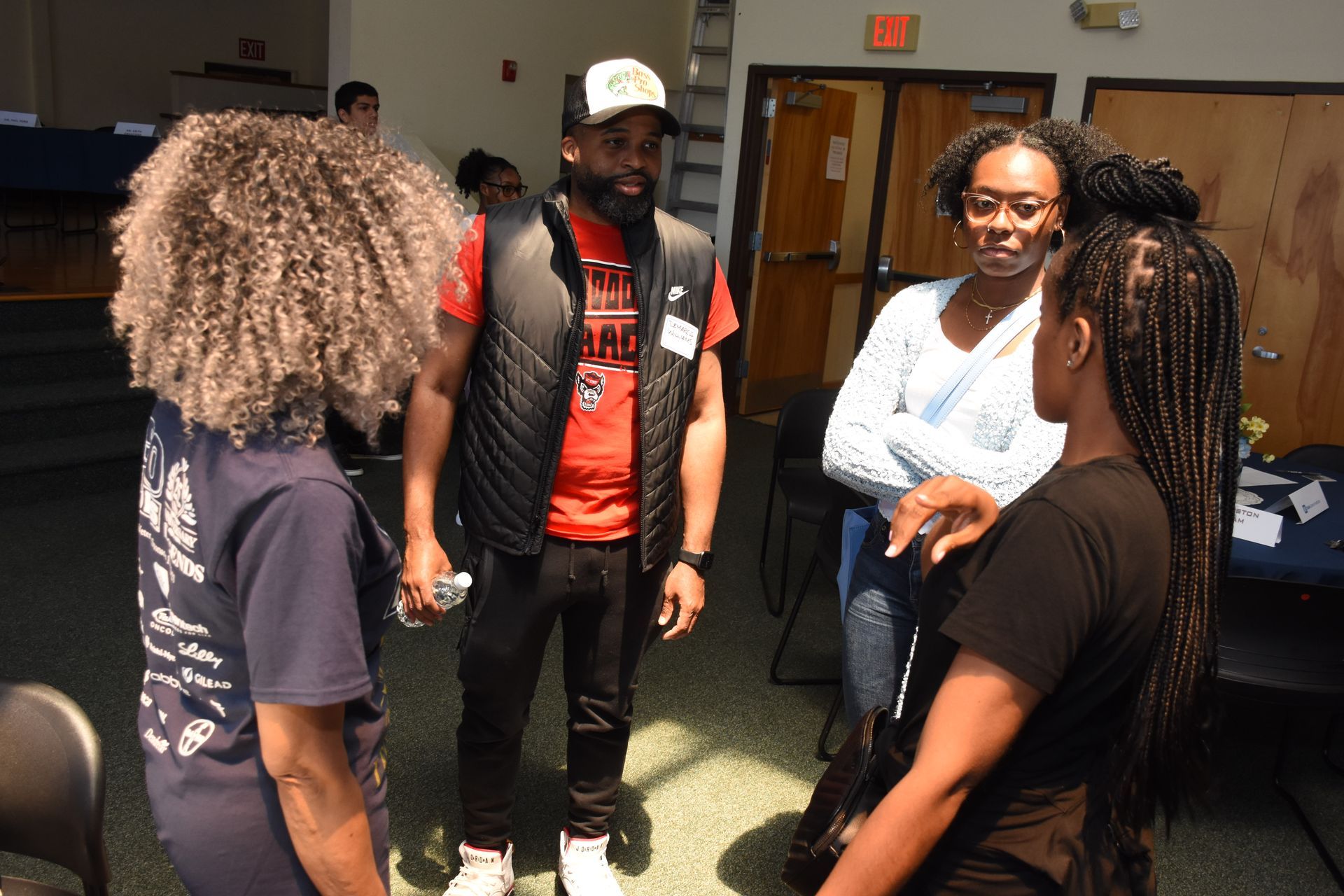 Four people in a room are in conversation. One man in a vest and hat is gesturing with his hands.