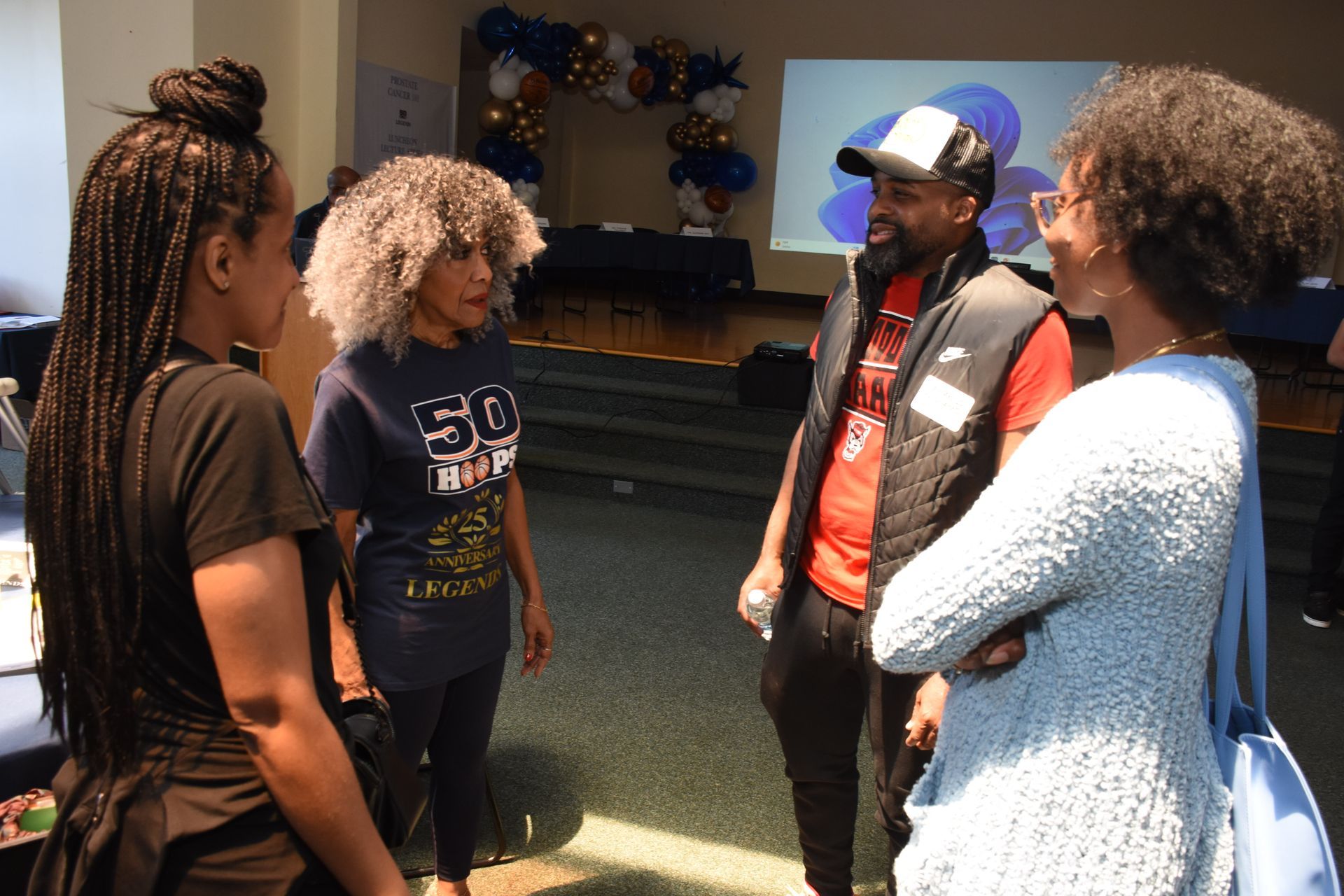 Four people talking in a room with balloons and a projection screen. One wears a black vest, another has grey hair.