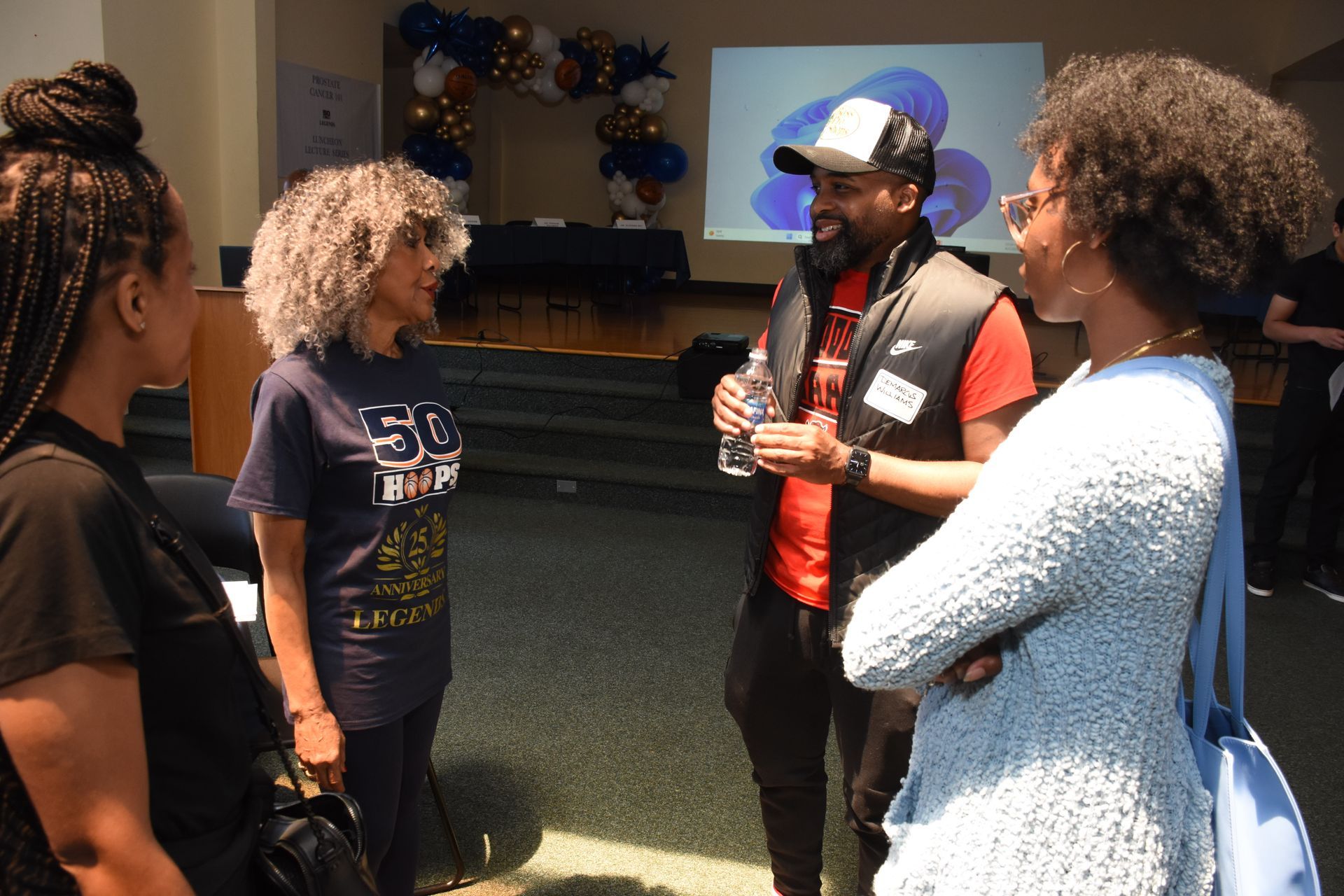 Four people conversing at an event. A man in a cap talks while others listen. Balloons and a screen are in the background.