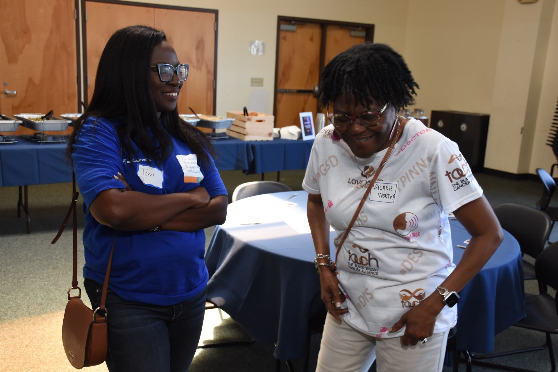 Two women talking at an event, one in blue, arms crossed; the other in white, looking down. Tables and doors in background.