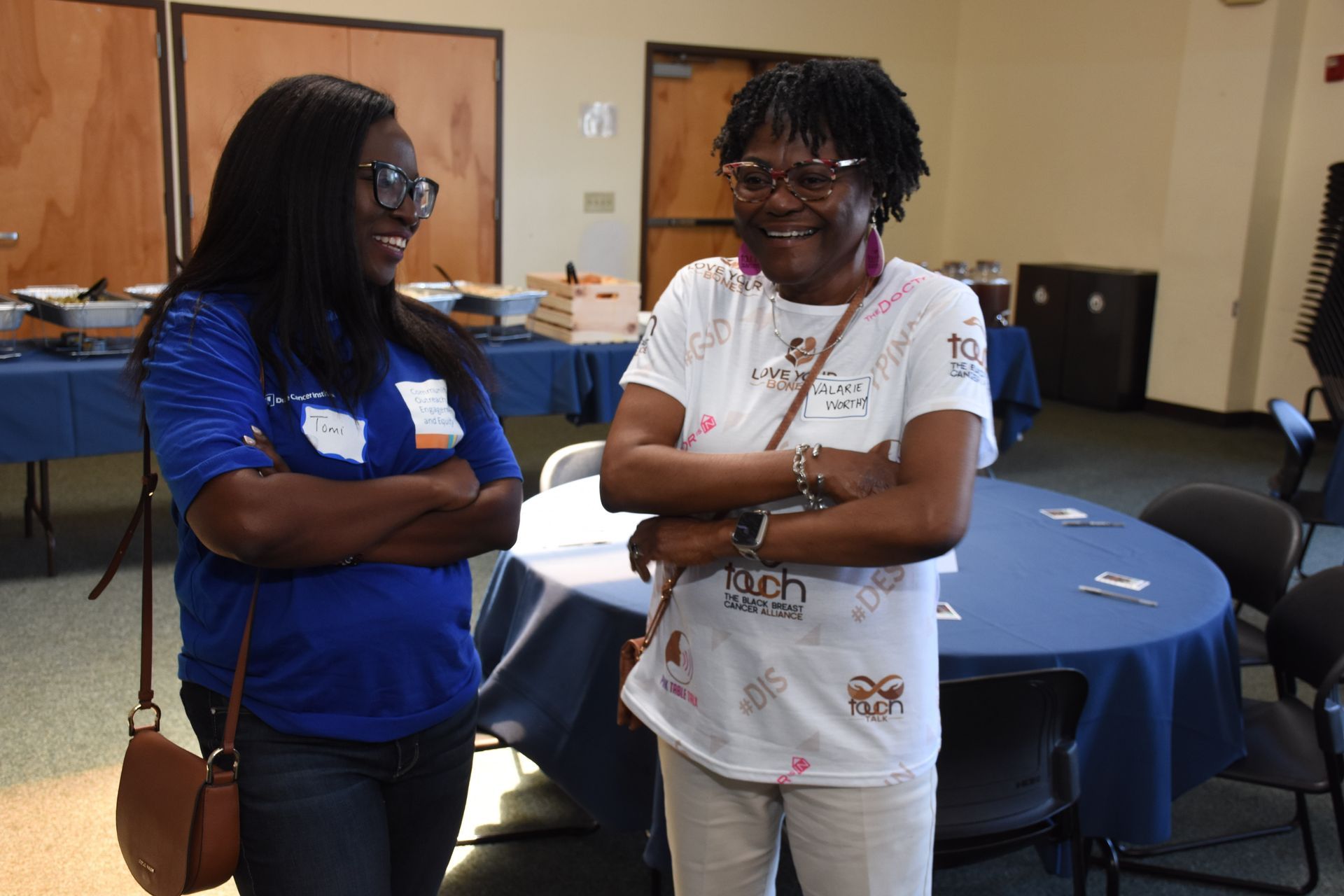 Two people, arms crossed, smiling, in a room with tables, blue tablecloths, and food.