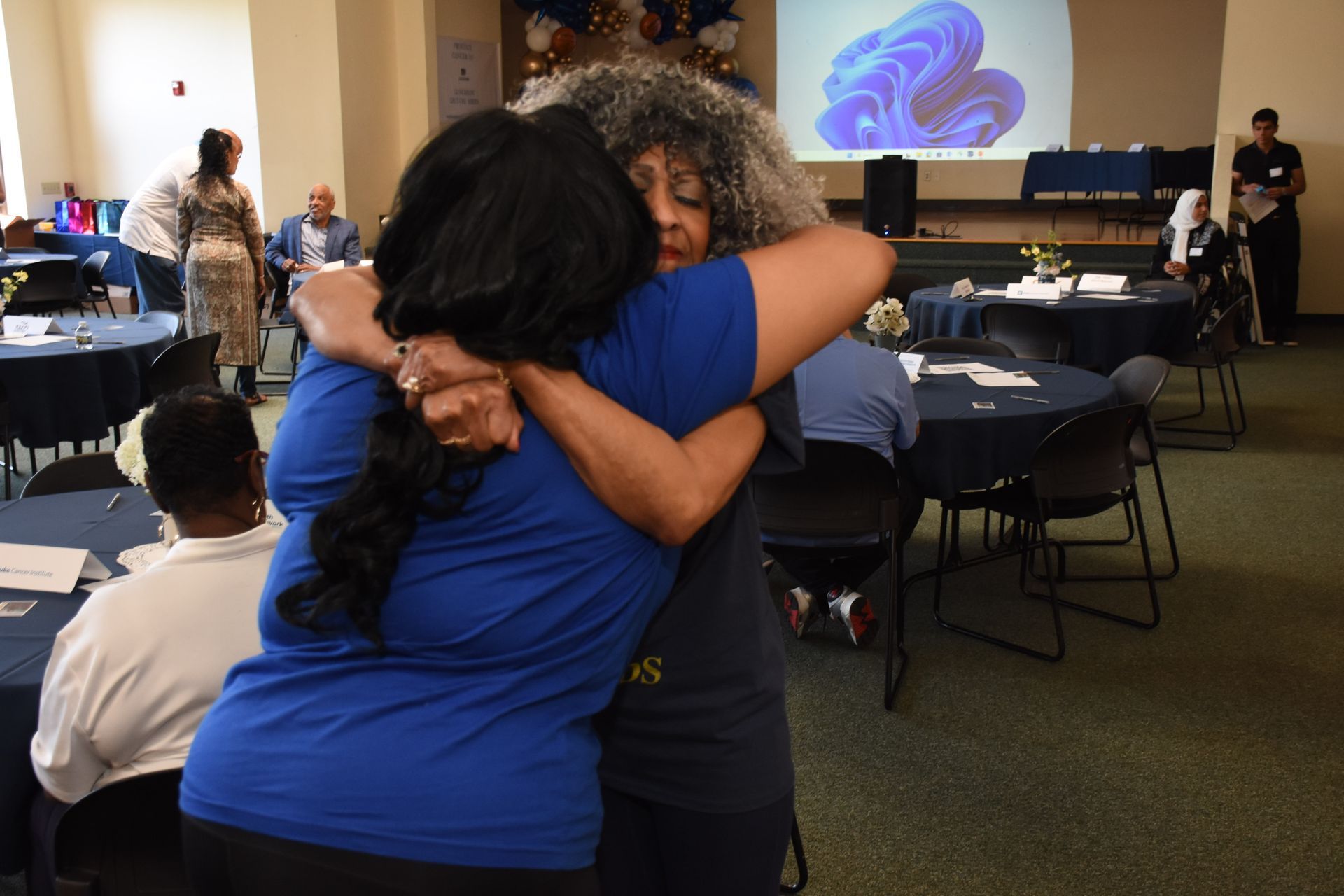 Two people embracing at an event with tables and a projection screen in the background.