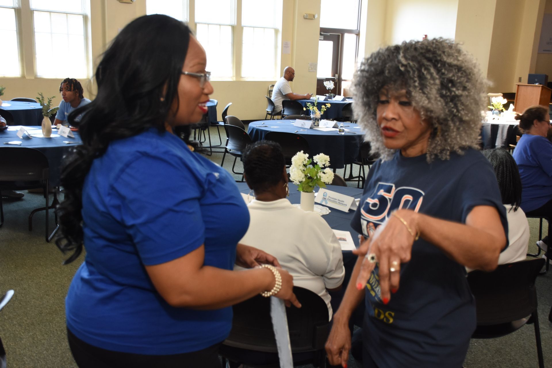Two women in blue shirts converse at a table-filled event. One has dark hair, the other has gray, curly hair.