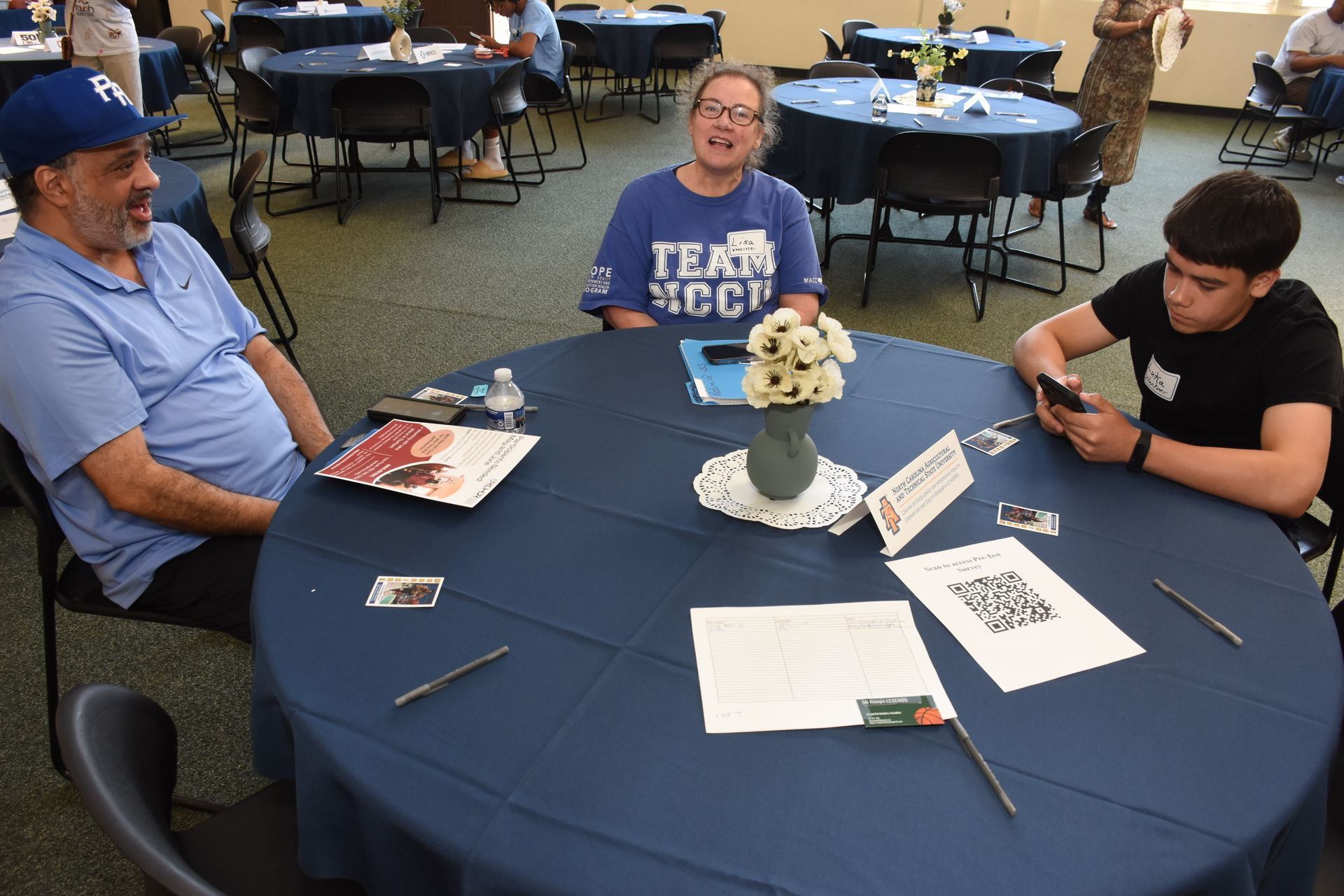 Three people seated at a round table, indoors. Two are smiling, one looks at their phone. Navy table cloth.