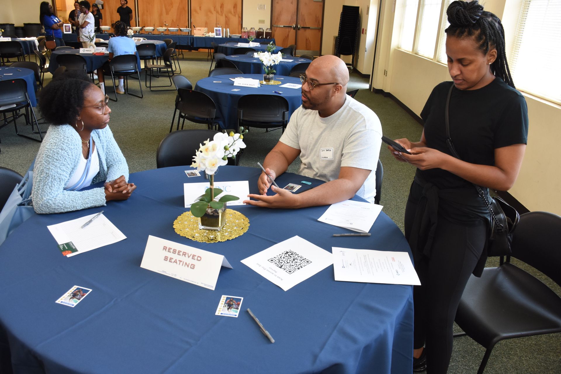 Three people at a table, one looking at phone. Event setting with blue tablecloths, name cards, and flower arrangement.