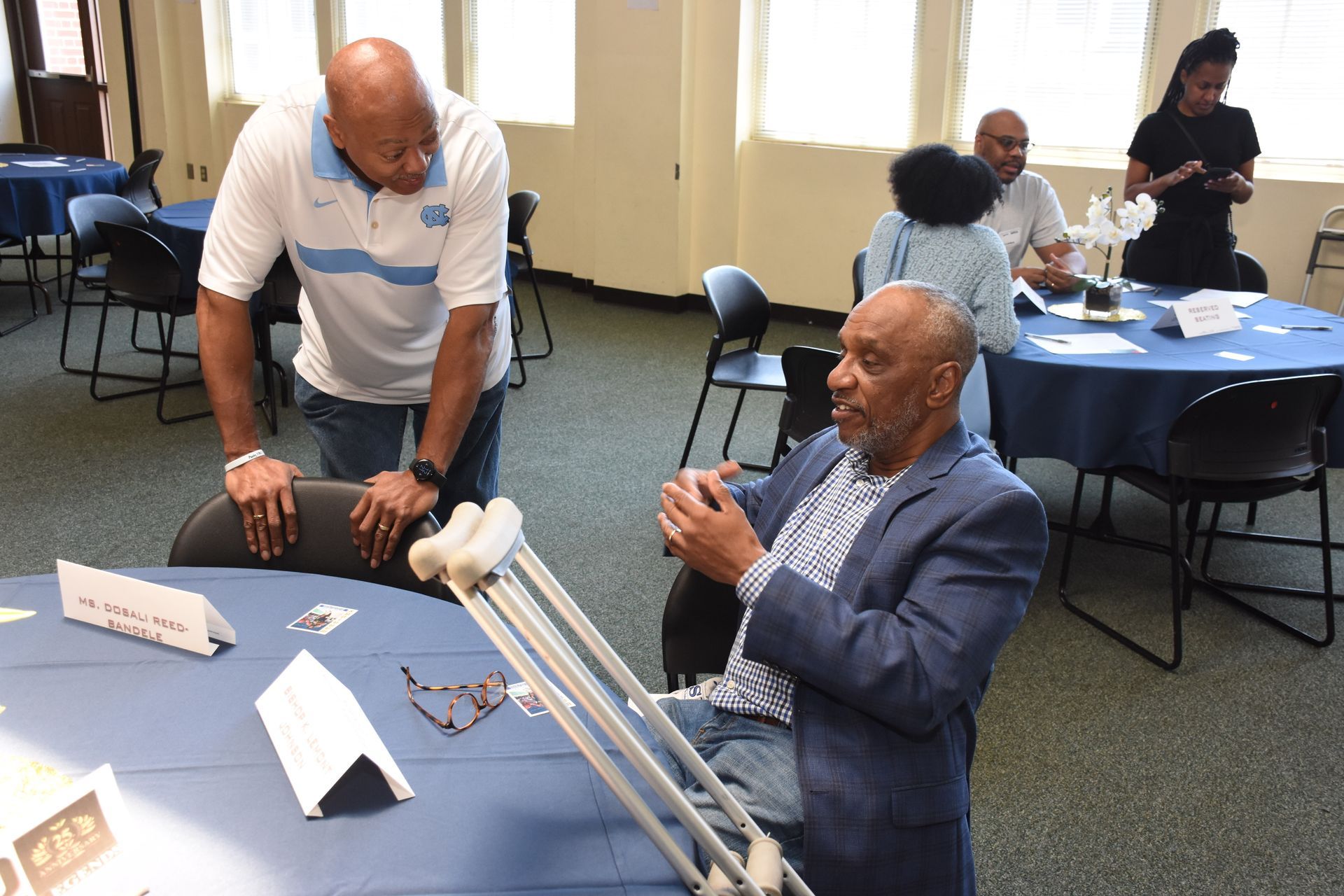 Man with crutches talking to another man in a room with round tables; other people visible.