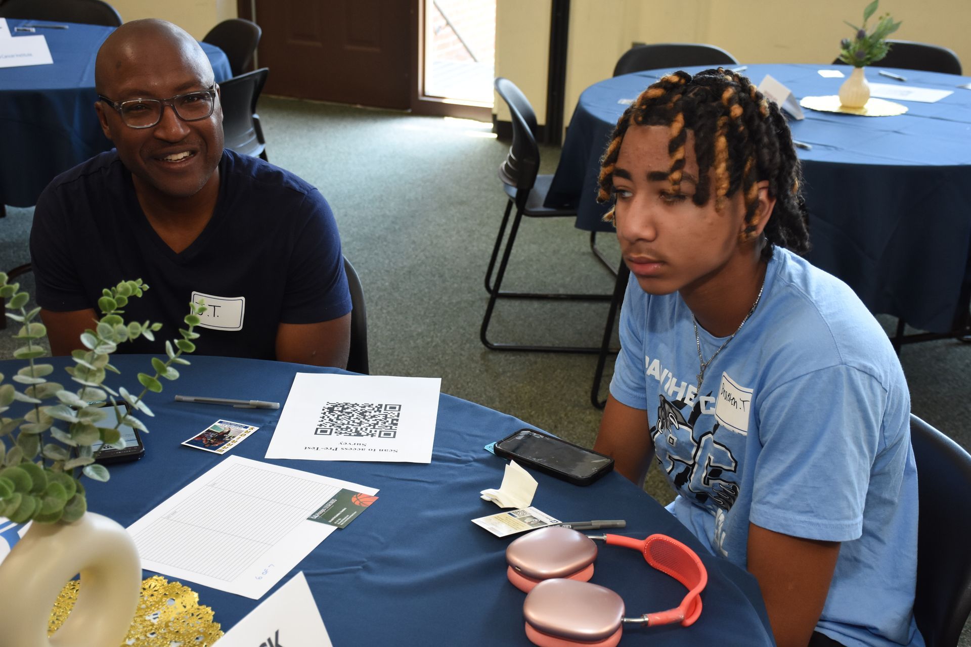 Two people at a table in a room with blue tablecloths. One has glasses, the other has dreadlocks and headphones.