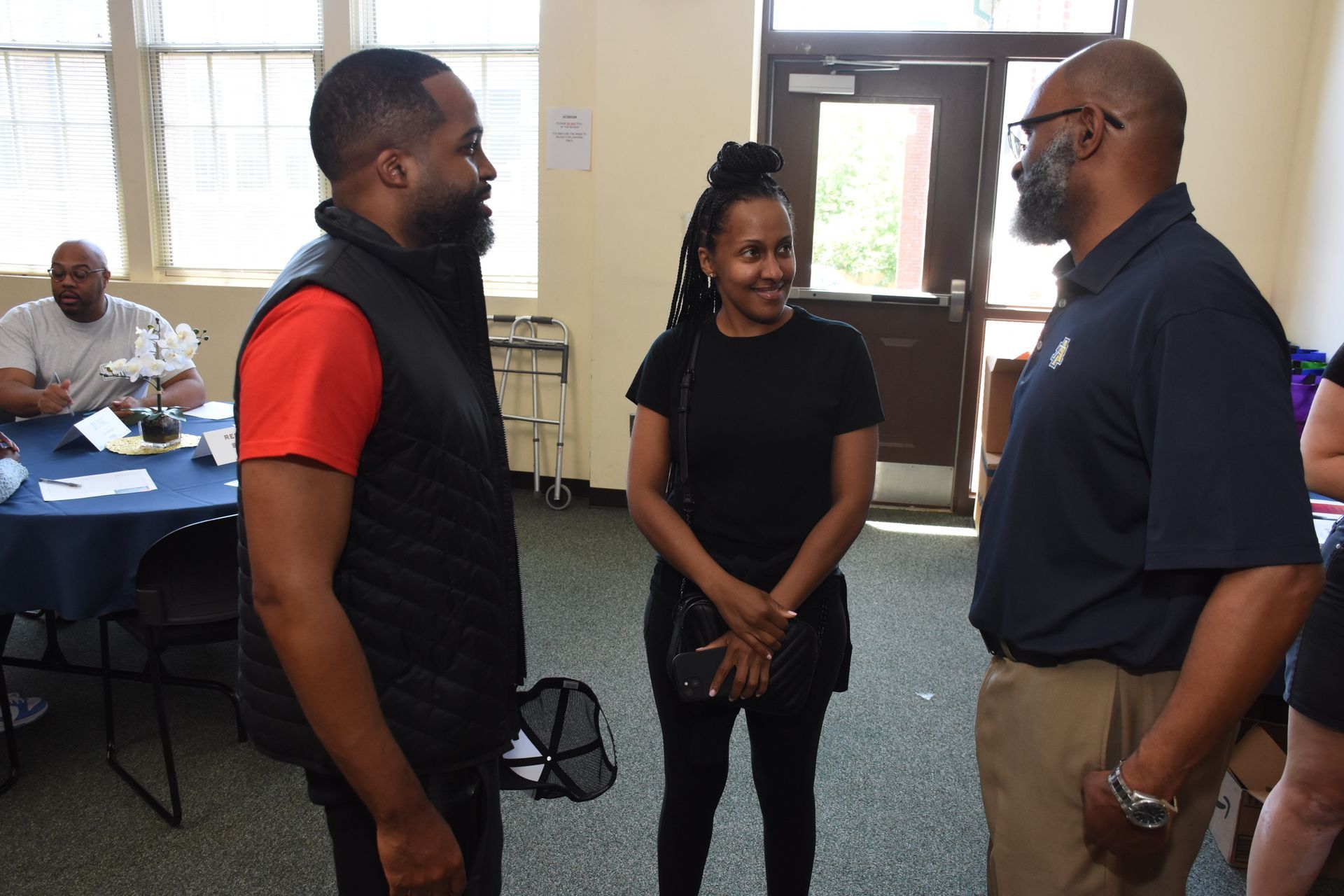 Three people talking in a brightly lit room with a table and open door in background.