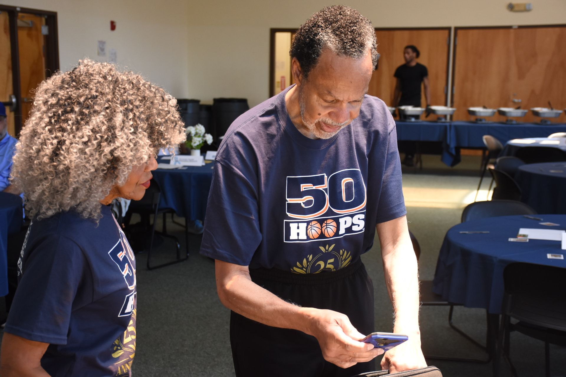 Two people looking at a phone in a room with tables and chairs. Both wear blue shirts with 