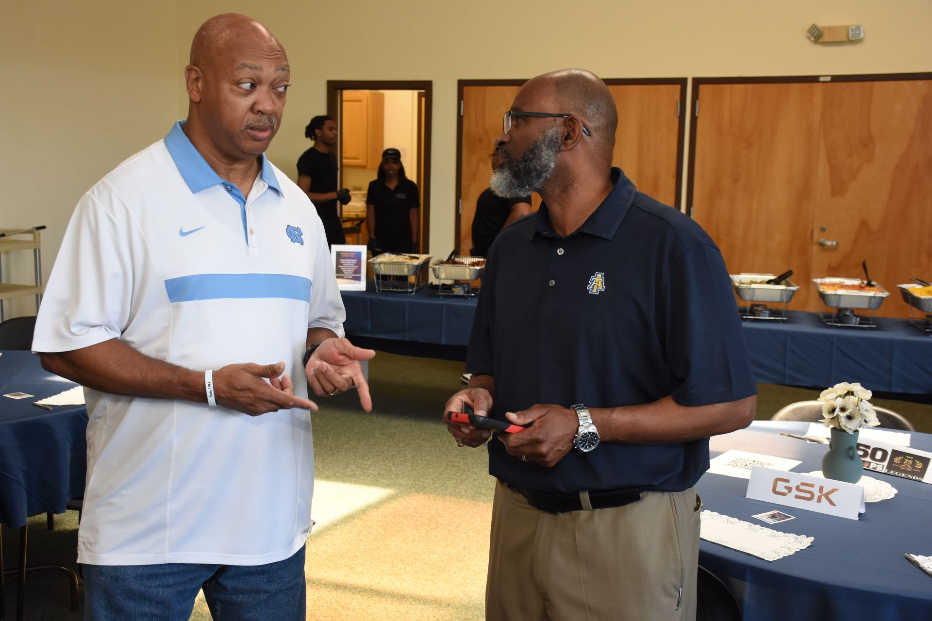 Two men conversing near a buffet table. The man on the left gestures while speaking; the other listens.