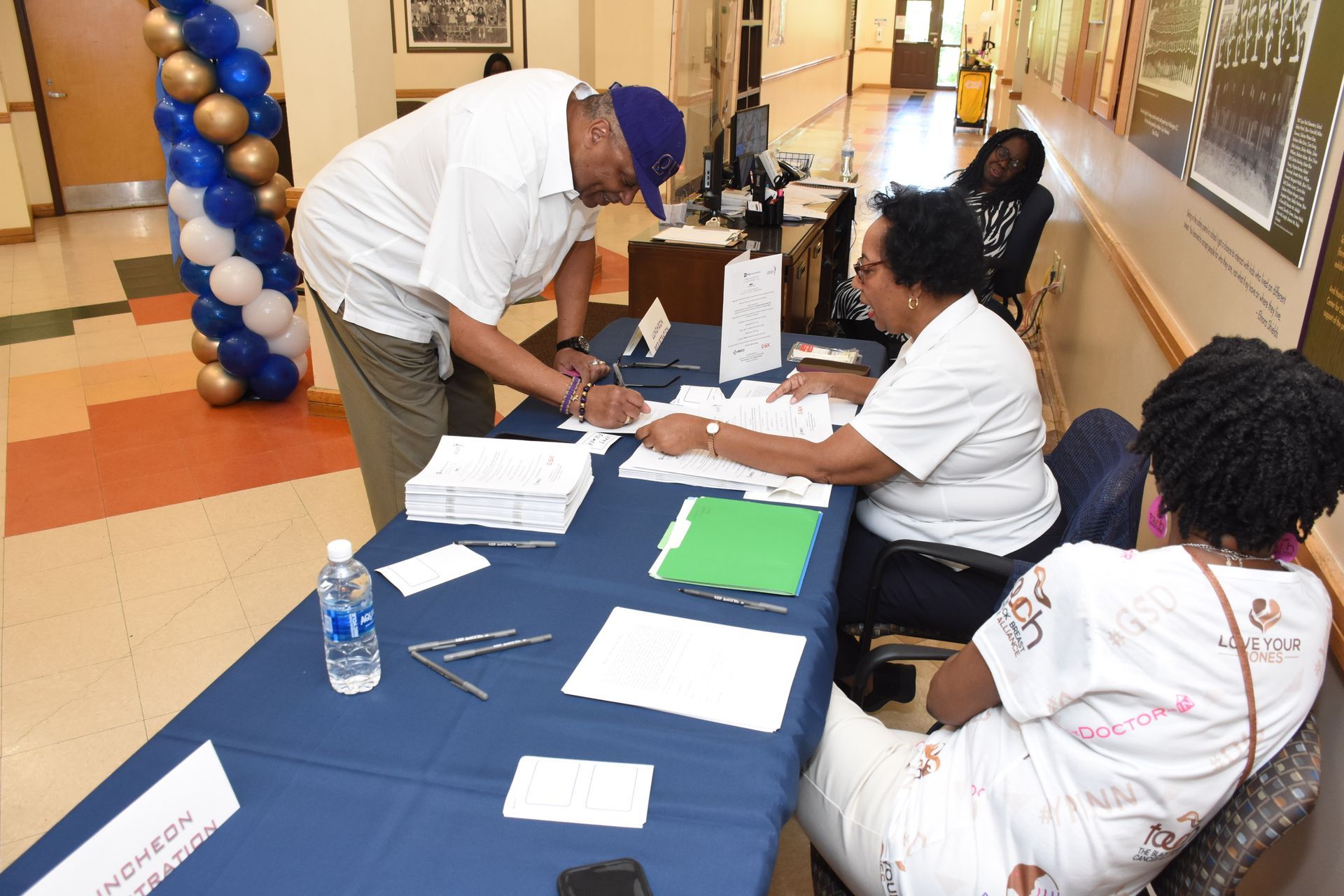 People signing forms at a table in a hall with a balloon arch.